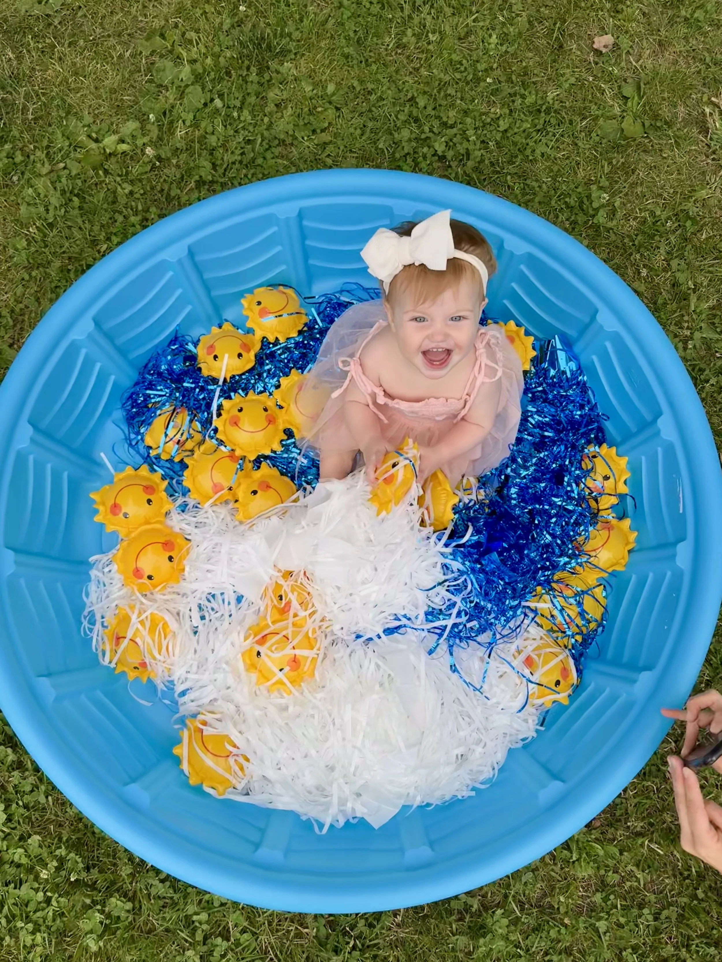 Young girl sitting in a blue plastic pool filled with yellow rubber duck toys, surrounded by blue and white shredded paper, on grass, smiling up at the camera.