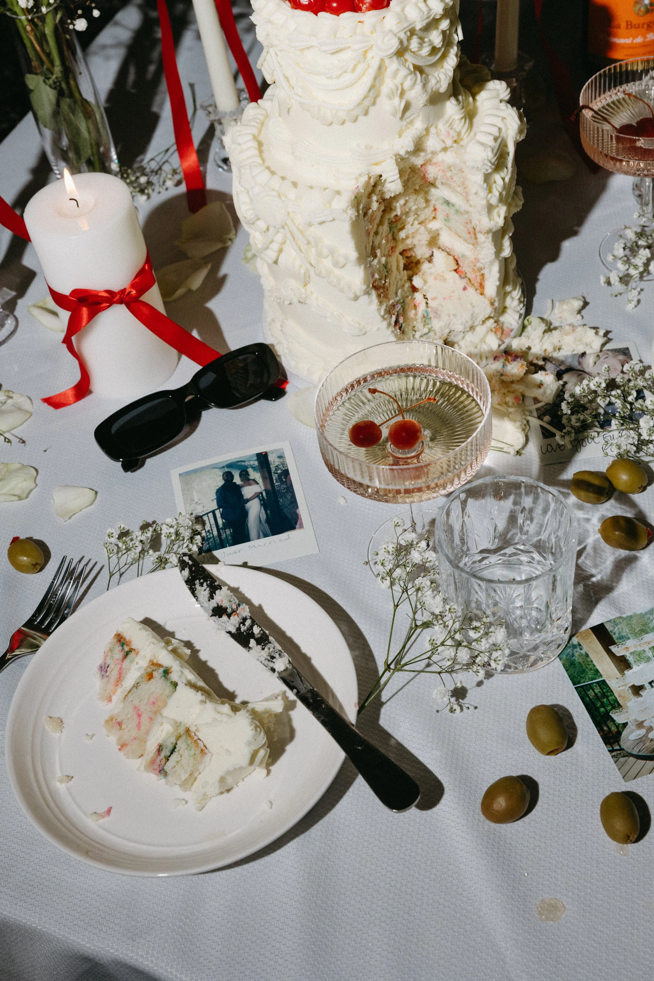 Partially eaten three-tiered white wedding cake with colorful sprinkles inside, located on a decorated table with flowers, sunglasses, a candle with a red ribbon, a glass of cherries, and a vintage photograph.