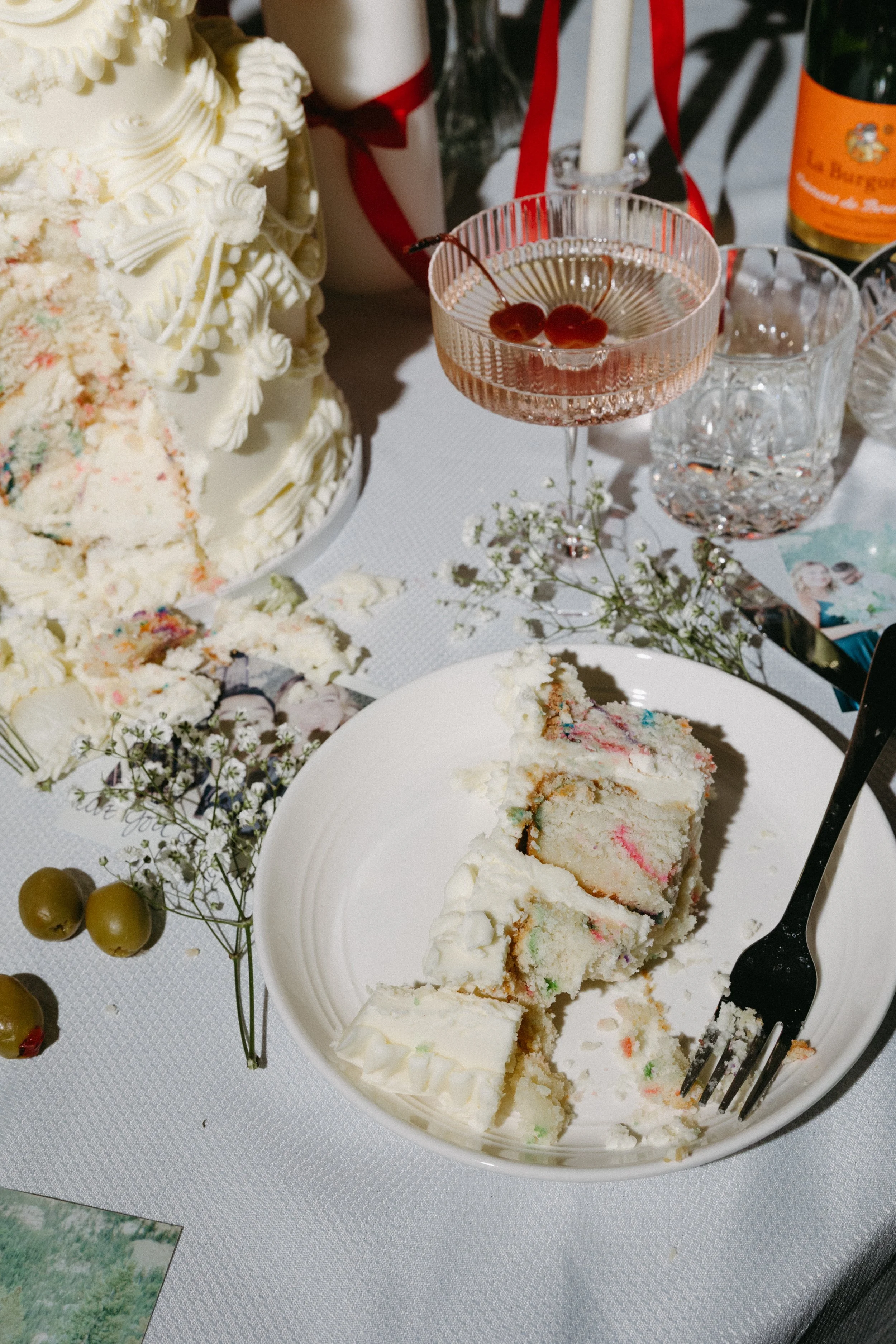 A slice of colorful cake on a white plate, surrounded by decorative flowers and green olives, on a table with a birthday cake, a cocktail glass with cherries, and various glasses and bottles.