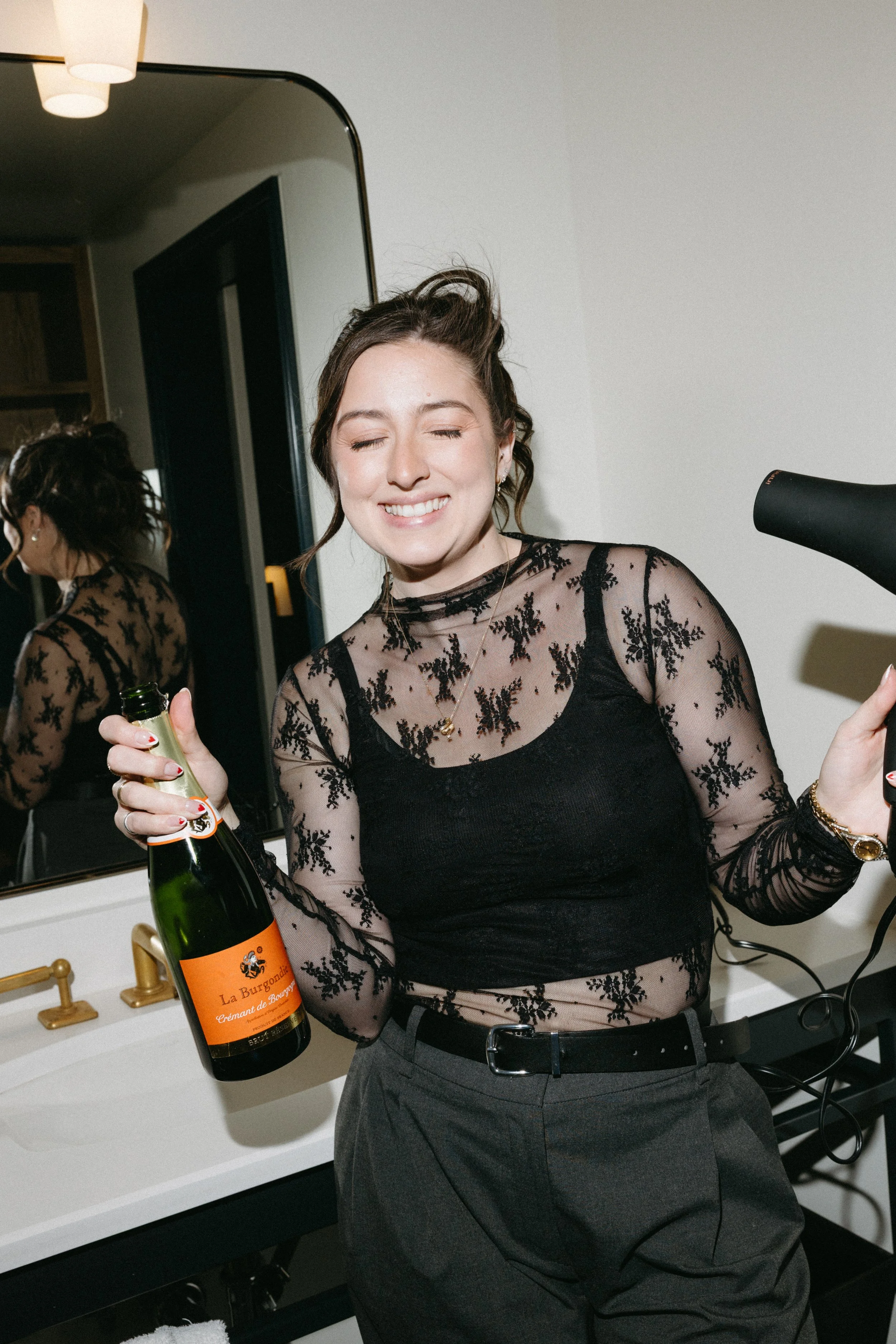 A young woman with dark hair styled in loose waves, smiling with eyes closed, holding a bottle of champagne in one hand and a hair dryer in the other. She is wearing a sheer black top with floral embroidery and dark pants, standing in a room with a mirror, sink, and gold faucet.
