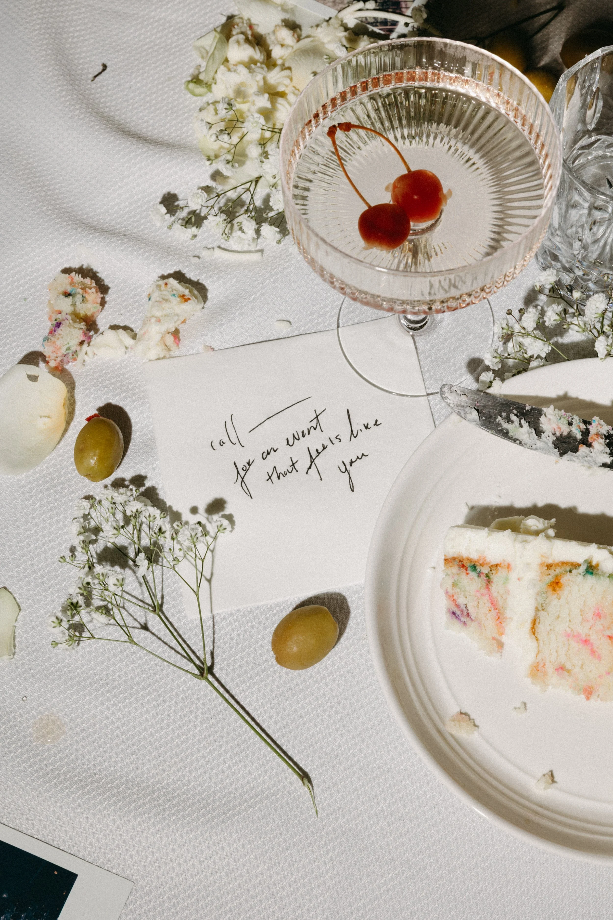 Partially eaten slice of colorful cake on a white plate, a glass of pink drink with cherries, flowers, grapes, and a handwritten note on paper, all on a white tablecloth.