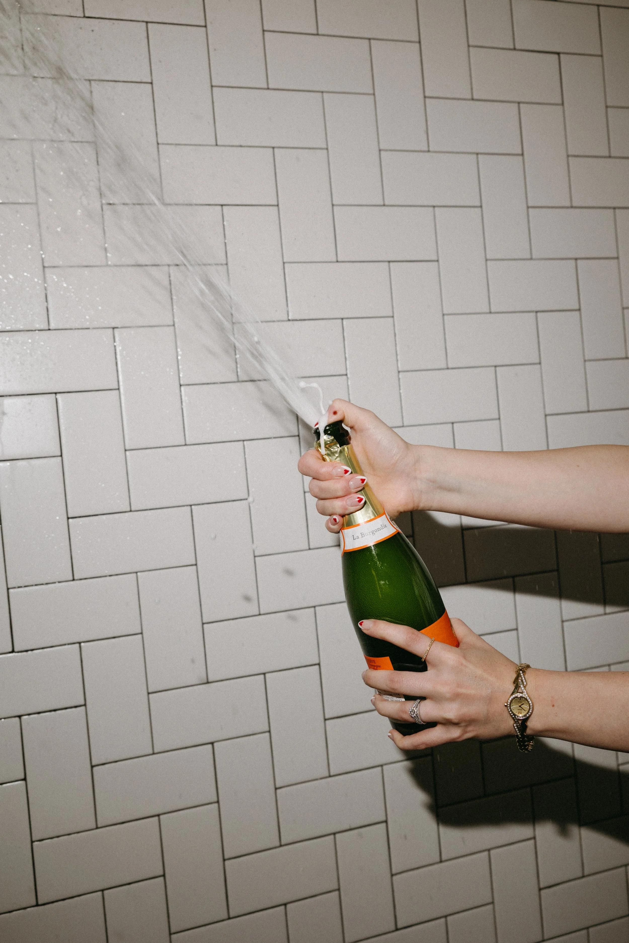 Person opening a bottle of champagne, with spray from the cork against a white tiled wall.