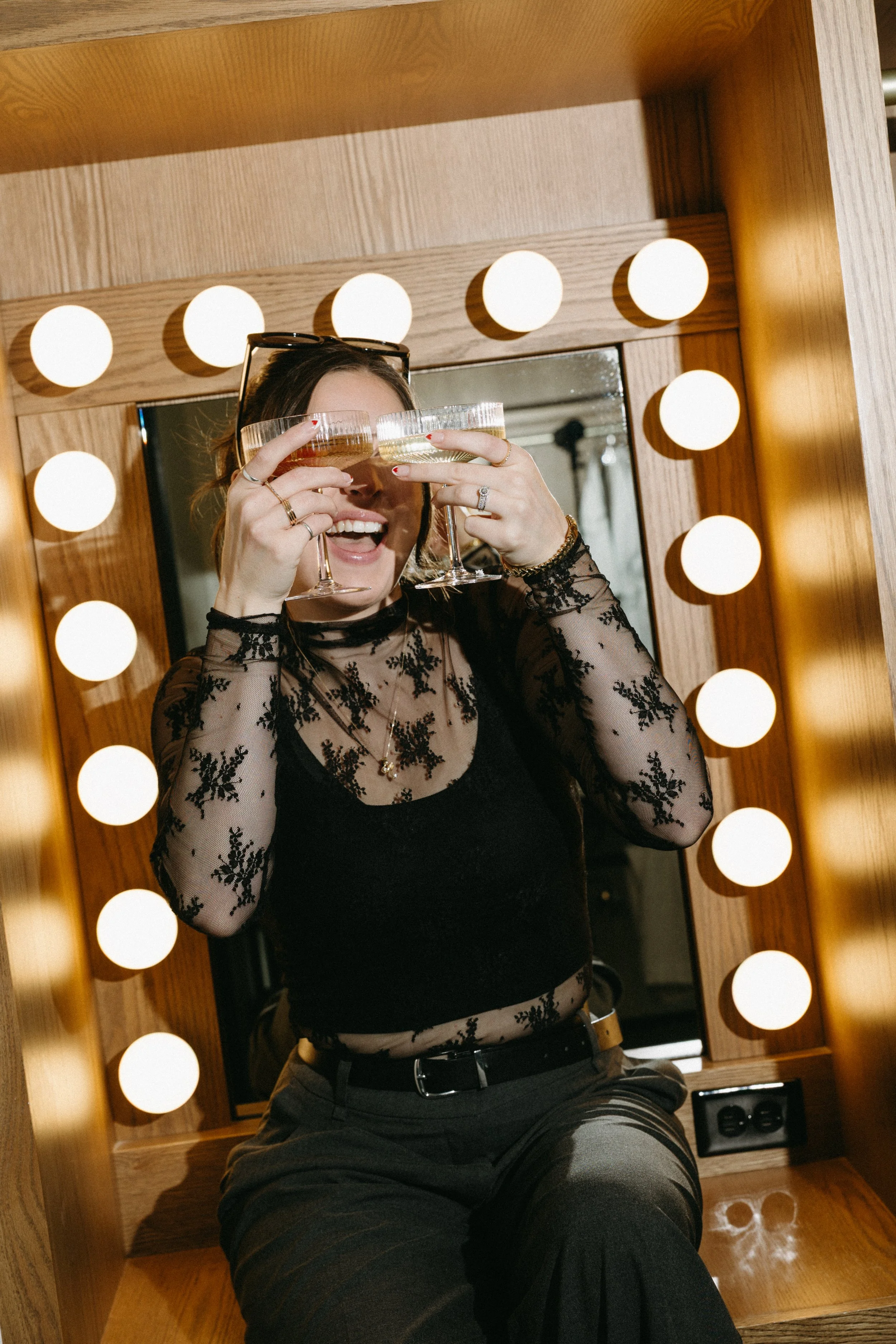 Woman in black lace top holding two champagne glasses, smiling, in front of a mirror surrounded by round light bulbs.