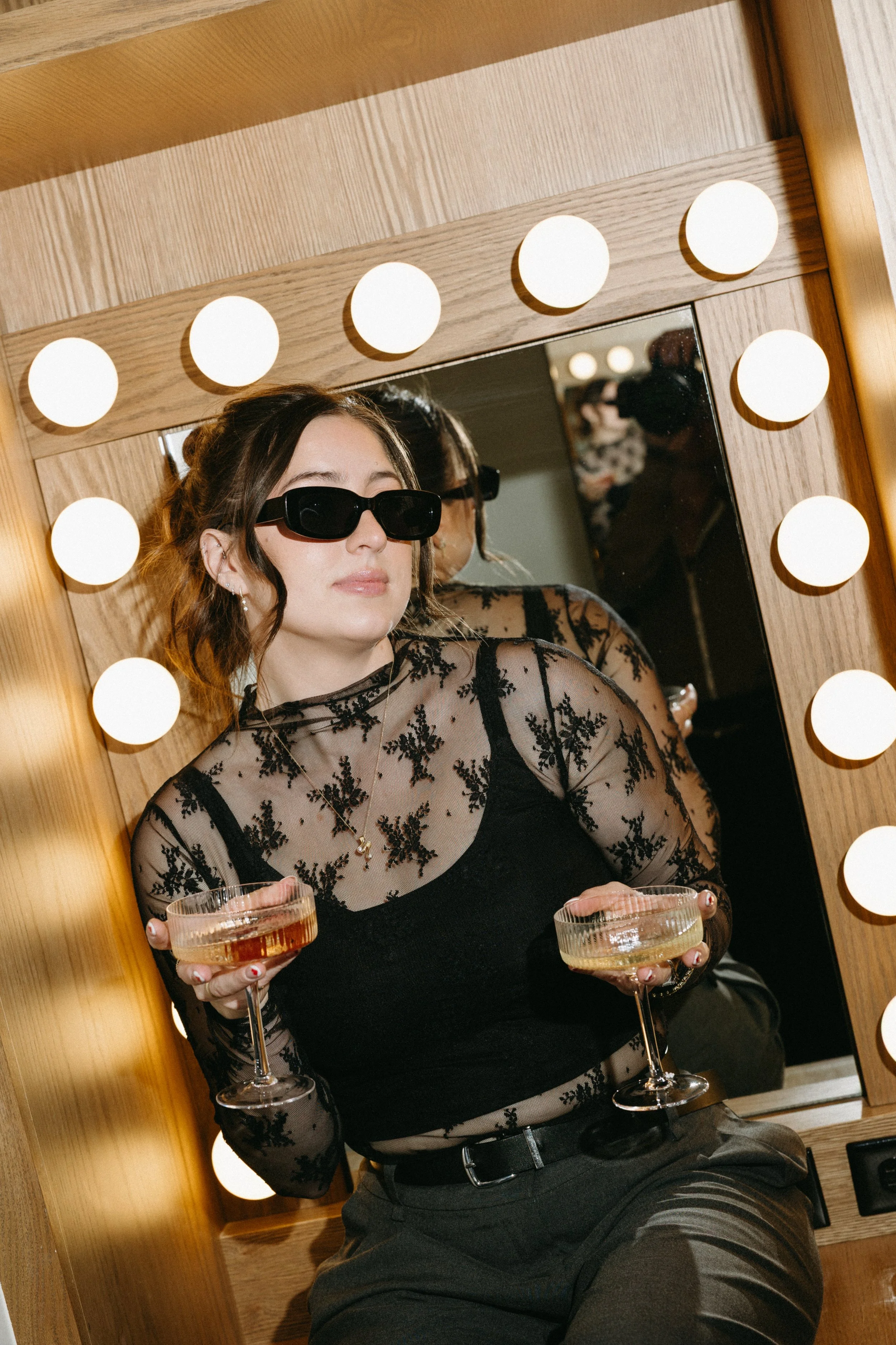 A woman with wavy brown hair wearing black sunglasses, a sheer black lace top with floral patterns, and dark pants, holding two cocktails in a backstage dressing room mirror with round bulb lights.