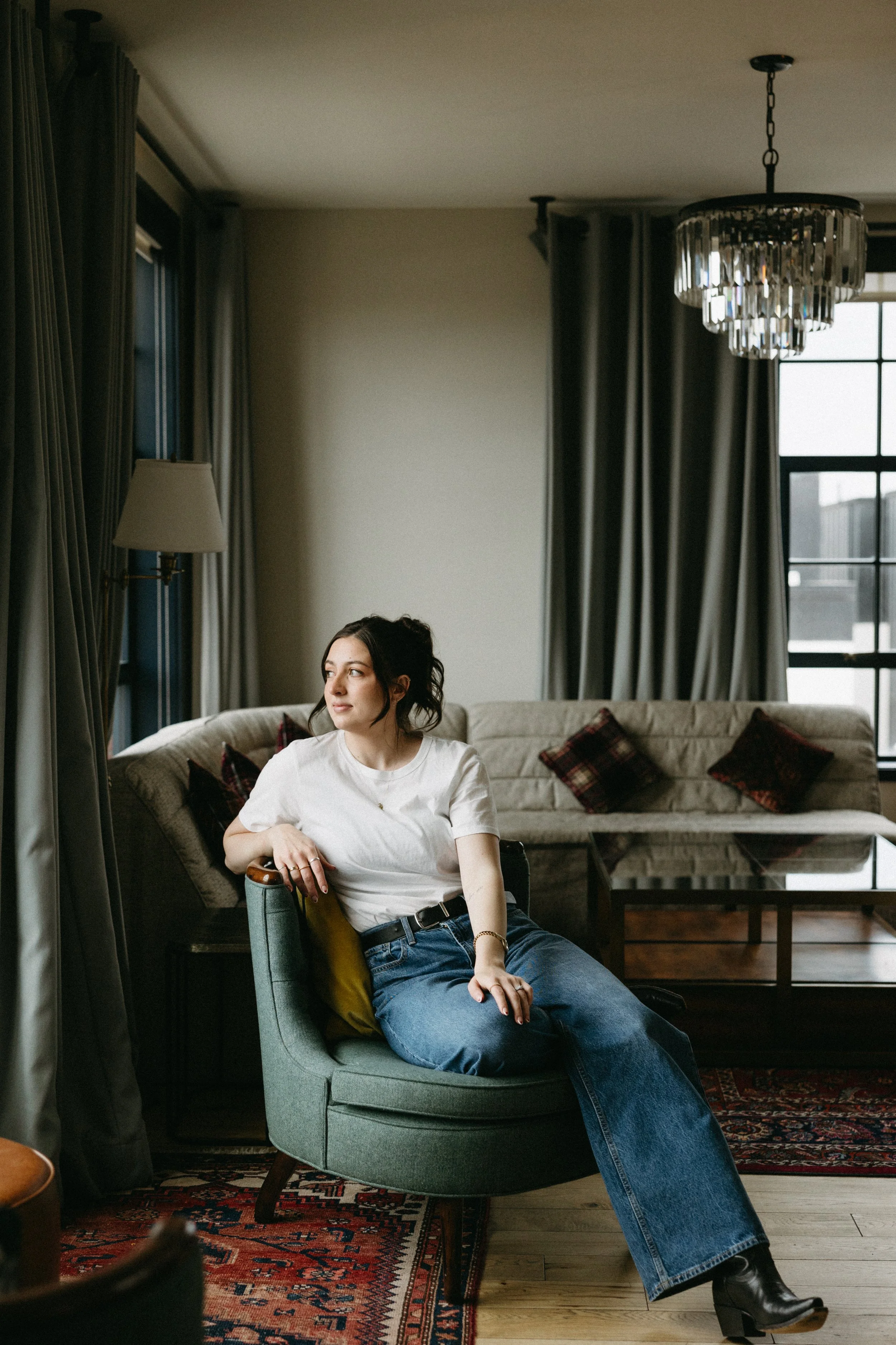 A woman with dark hair in a casual white t-shirt and blue jeans sitting on a green armchair in a cozy living room, looking thoughtfully out of a window towards the left.