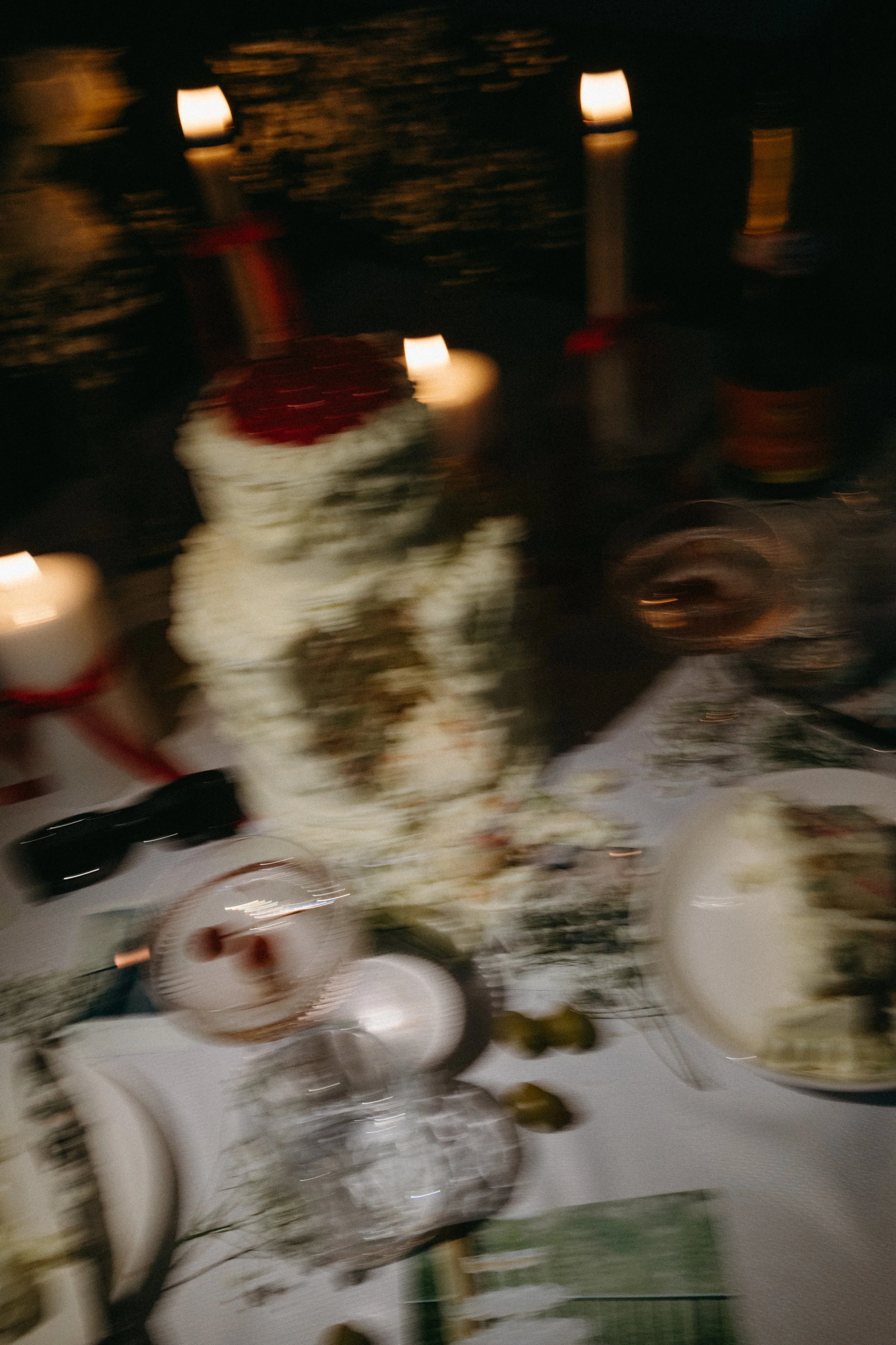 Blurred photo of a table with a wedding cake, candles, wine glasses, and dishes, likely at a celebration or wedding reception at night.
