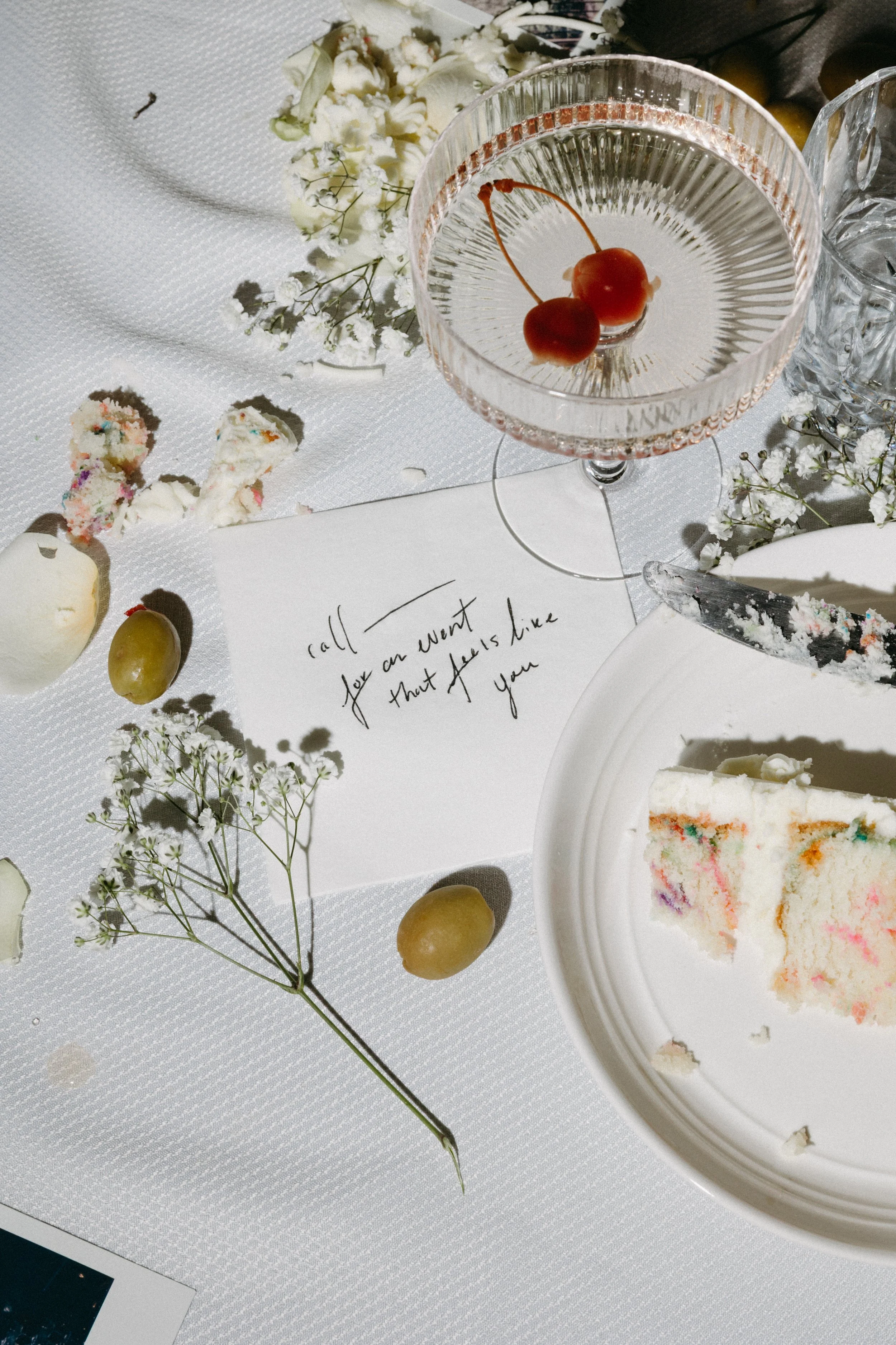 A partly eaten slice of rainbow cake on a white plate, a glass of cherry cocktail with two cherries, scattered green grapes, white flowers, and a note with handwritten text on a white tablecloth.