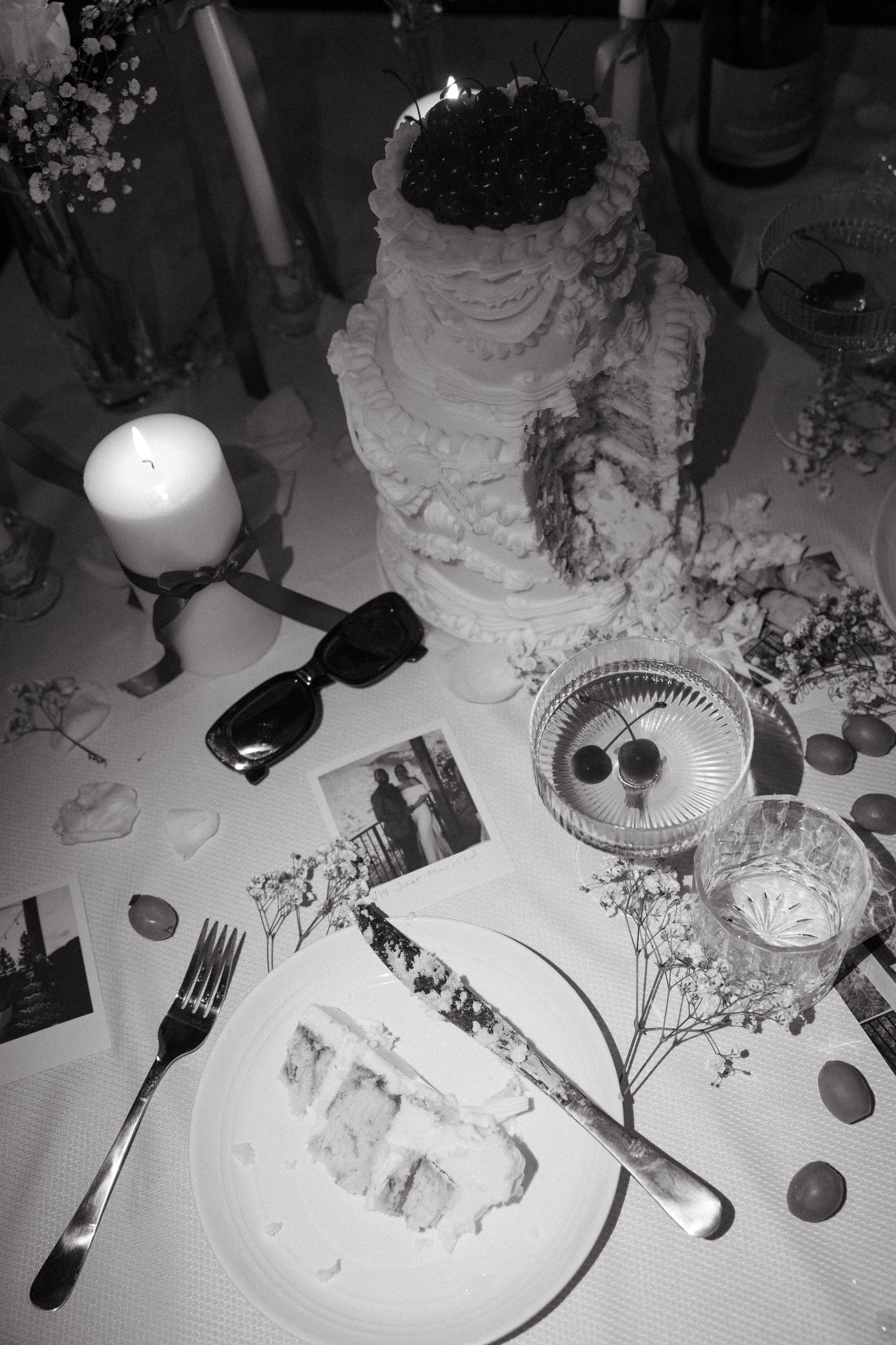 A partially eaten wedding cake, a lit candle, a pair of sunglasses, a photograph, and drinks with cherries on a decorated table.