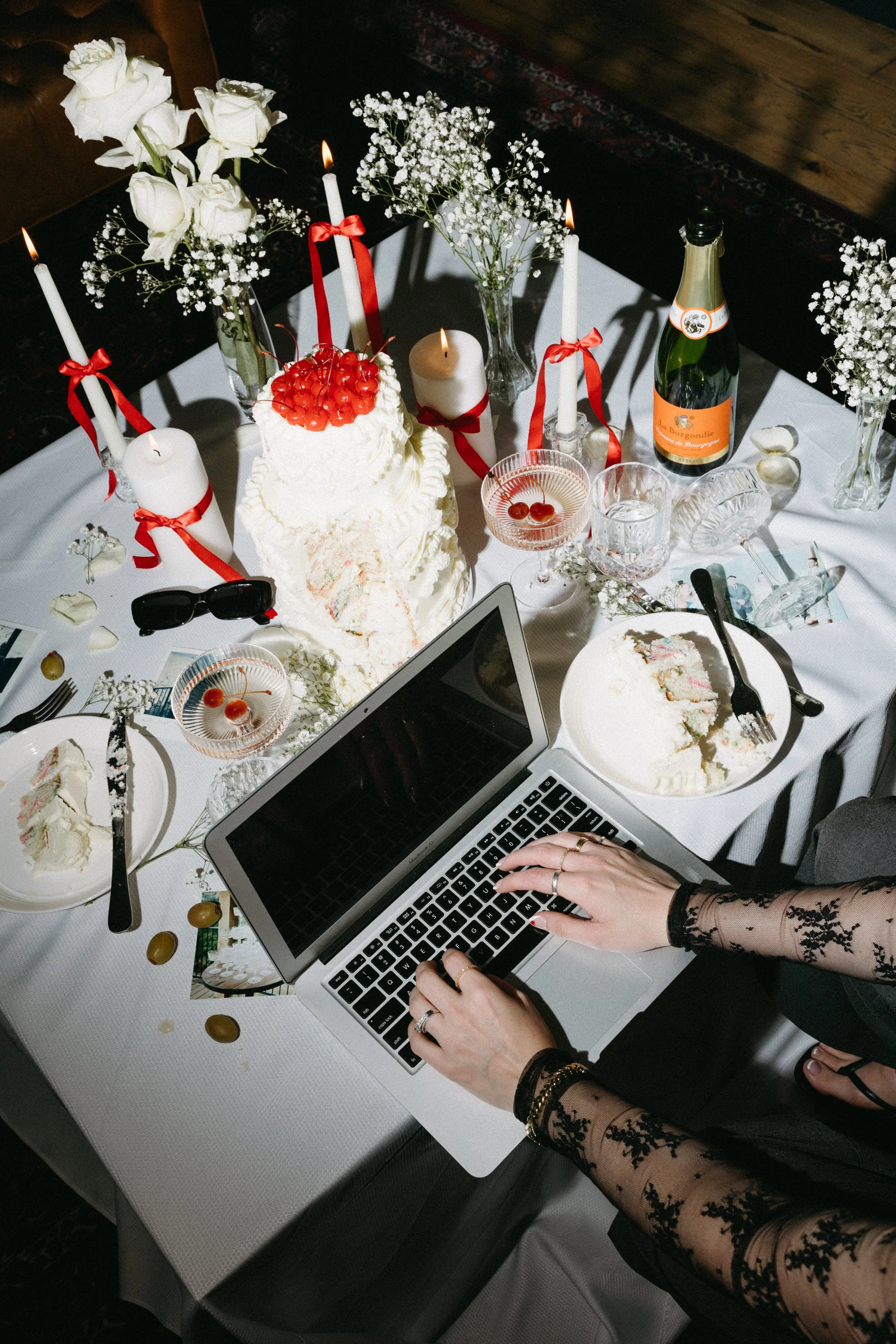 A birthday celebration table with a partially eaten cake, candles, flowers, champagne, and a person working on a laptop.