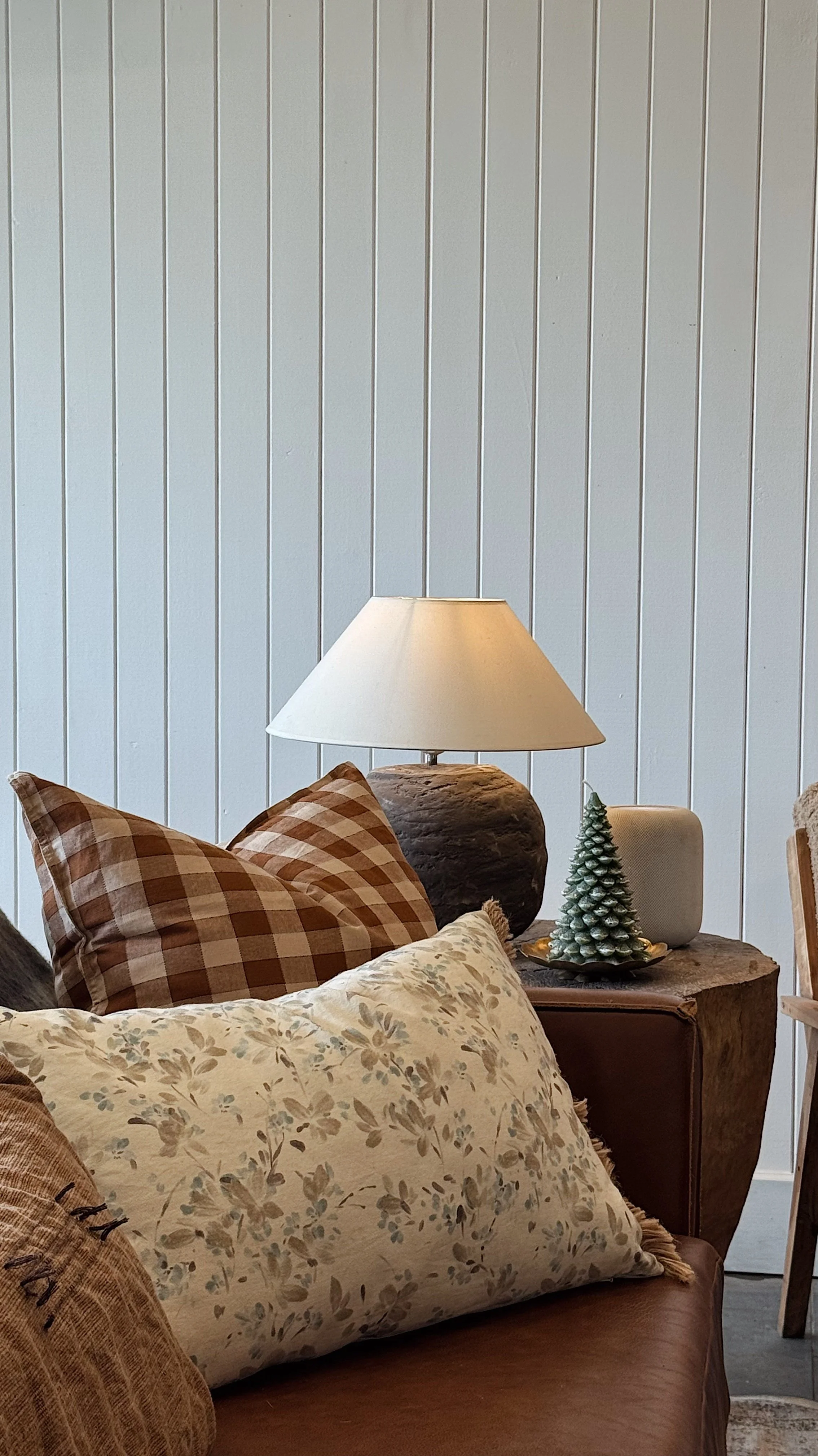 Living room corner with a couch, floral and checkered pillows, a wooden side table with a lamp, a tiny green Christmas tree, and a small white speaker against a white wooden-paneled wall.