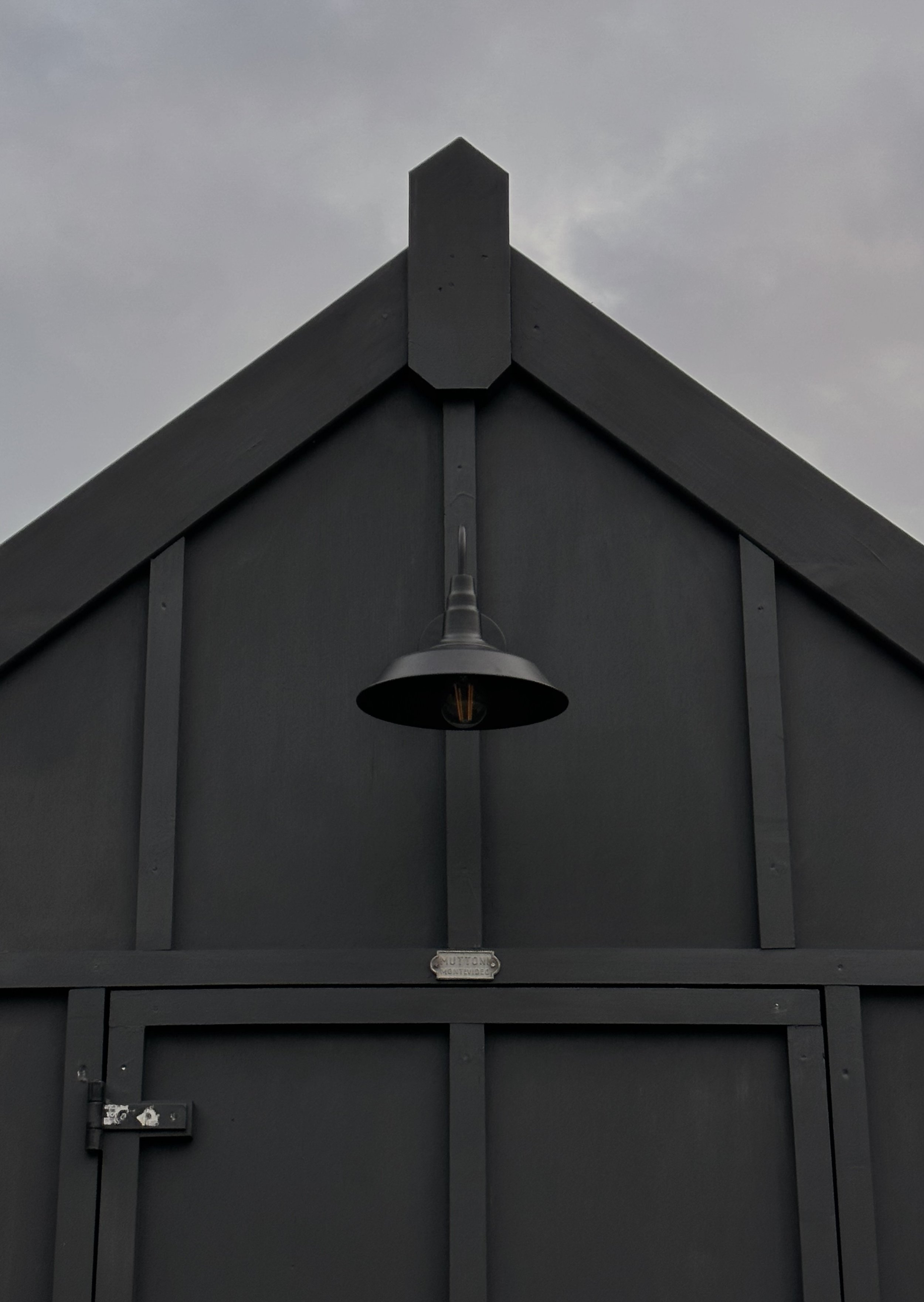 Estructura de una casa o edificio de color gris con una lámpara colgante en la fachada y un cielo nublado de fondo.