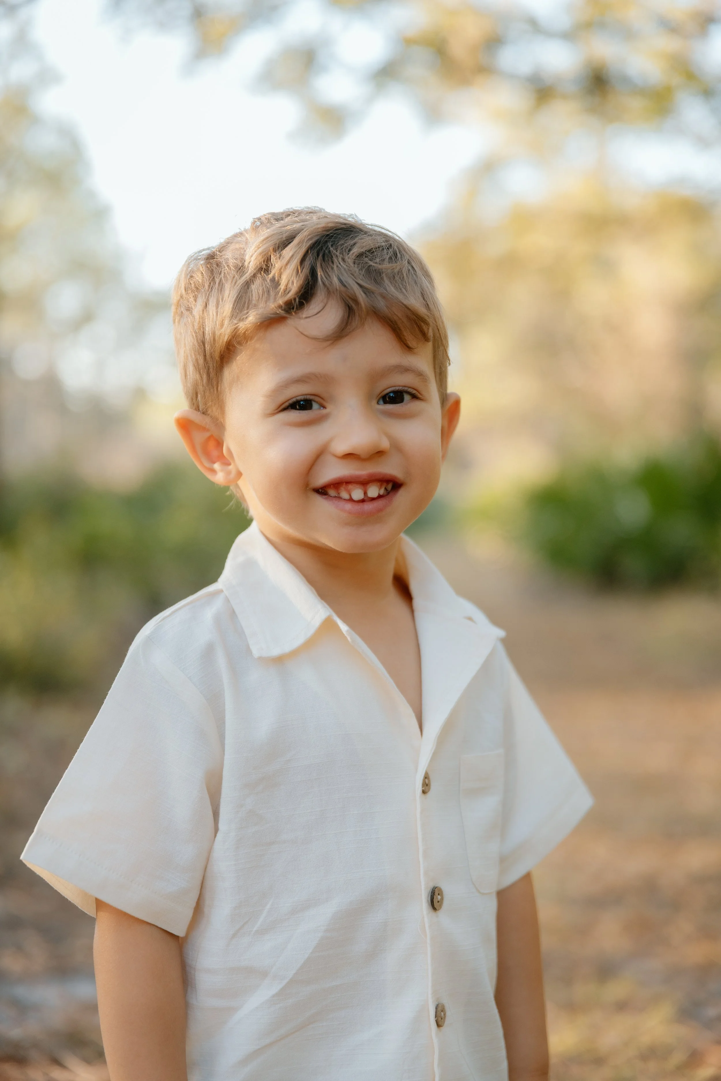 A young boy with light brown hair in a white button-up shirt, smiling outdoors in a park with trees and sunlight in the background.