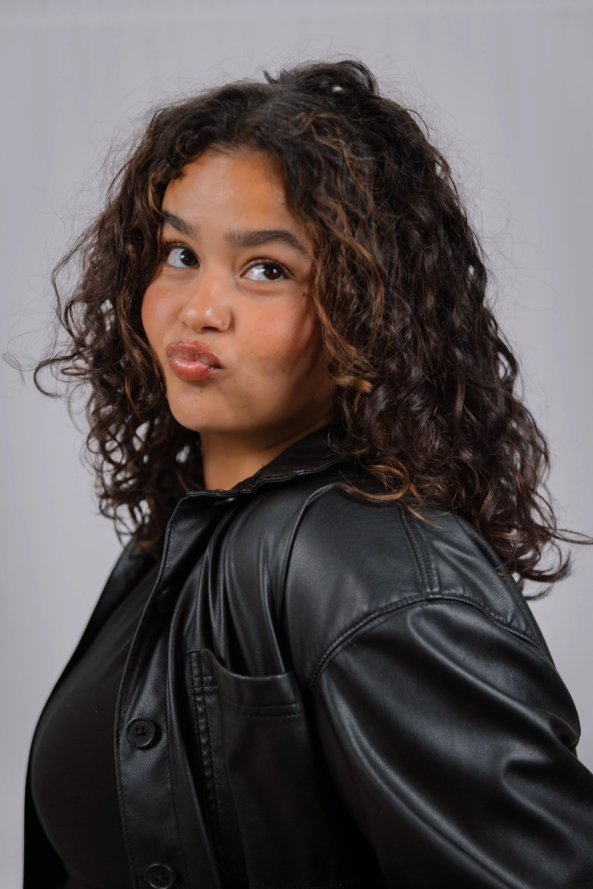 A woman with curly dark hair making a pouty face, wearing a black leather jacket, against a plain light grey background.