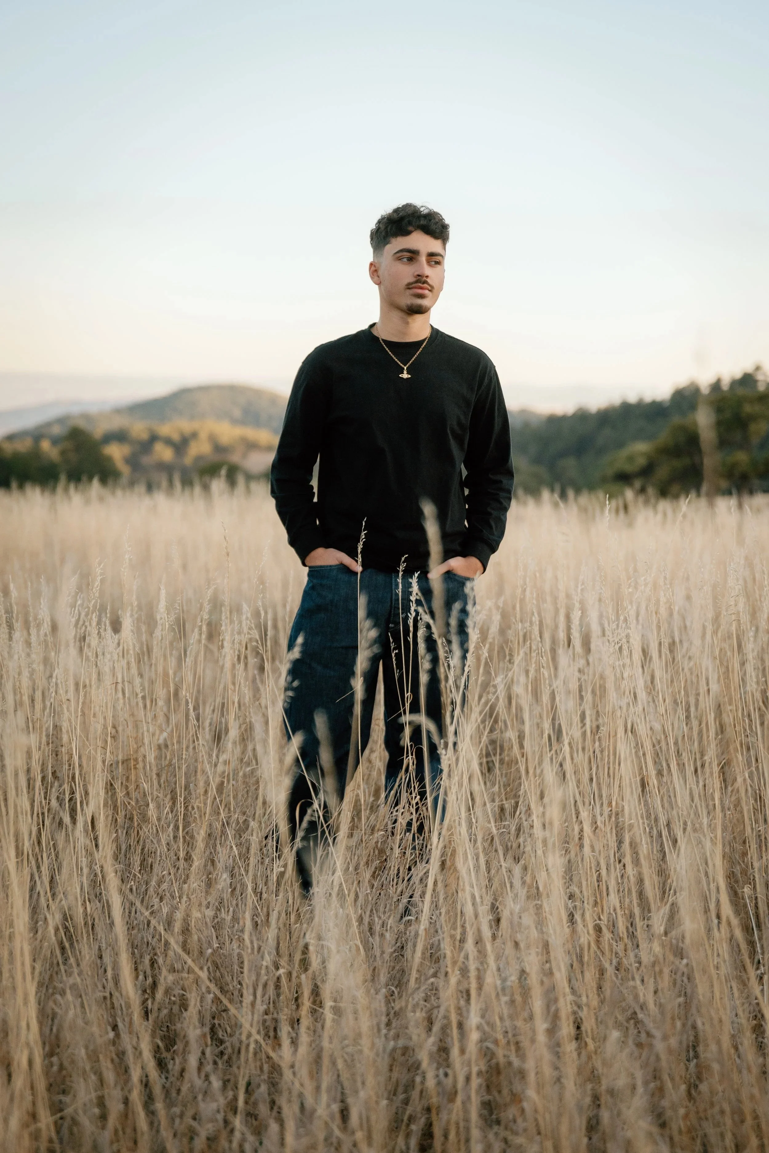 A young man standing in a field of tall dry grass with mountains and a pale sky in the background.