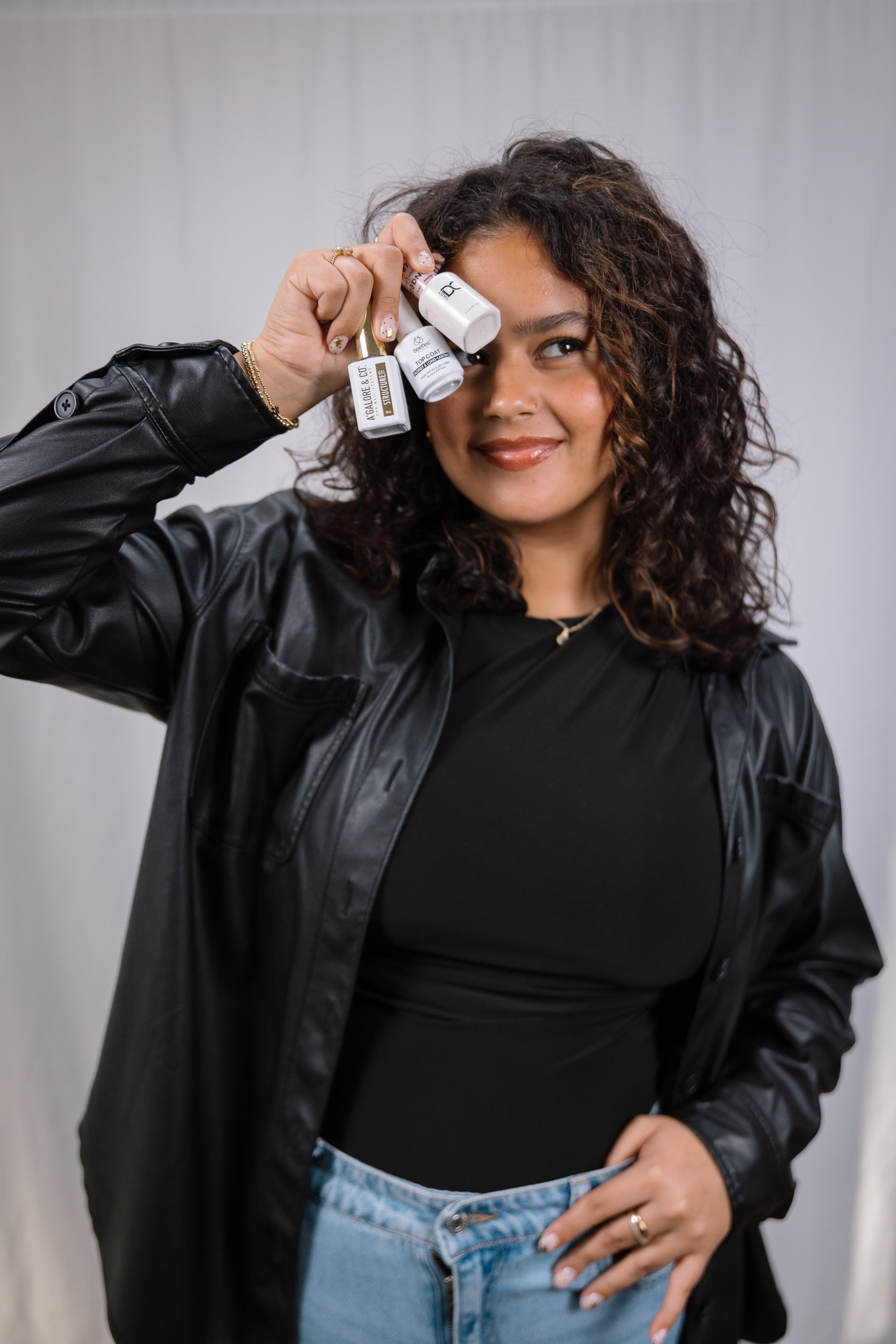 Woman with dark curly hair wearing a black leather jacket and black top, holding skincare products near her forehead, smiling, standing in front of a plain background.
