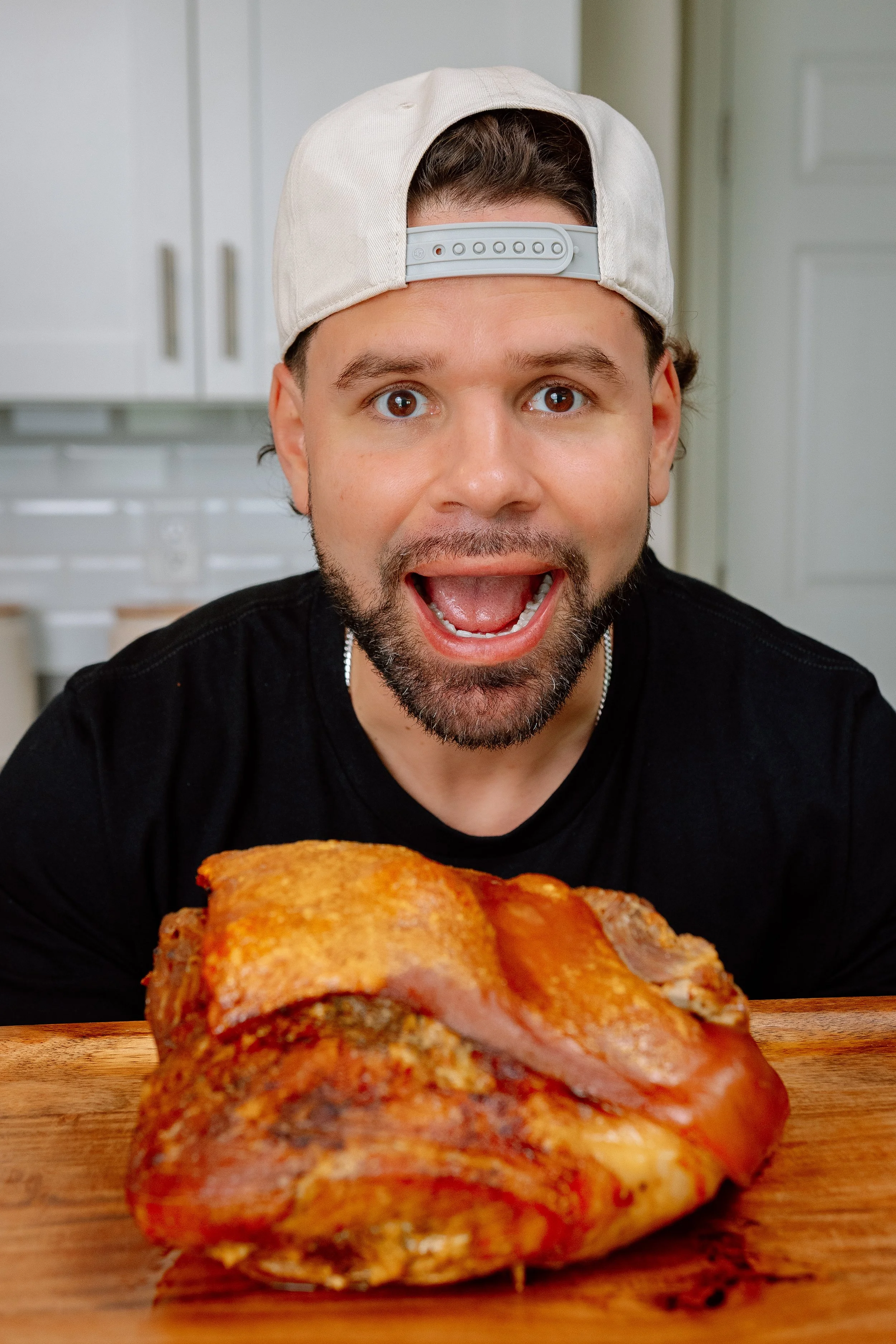 A man with a beard and a baseball cap backwards, smiling with an open mouth, leaning over a large piece of raw pork on a wooden cutting board in a kitchen.