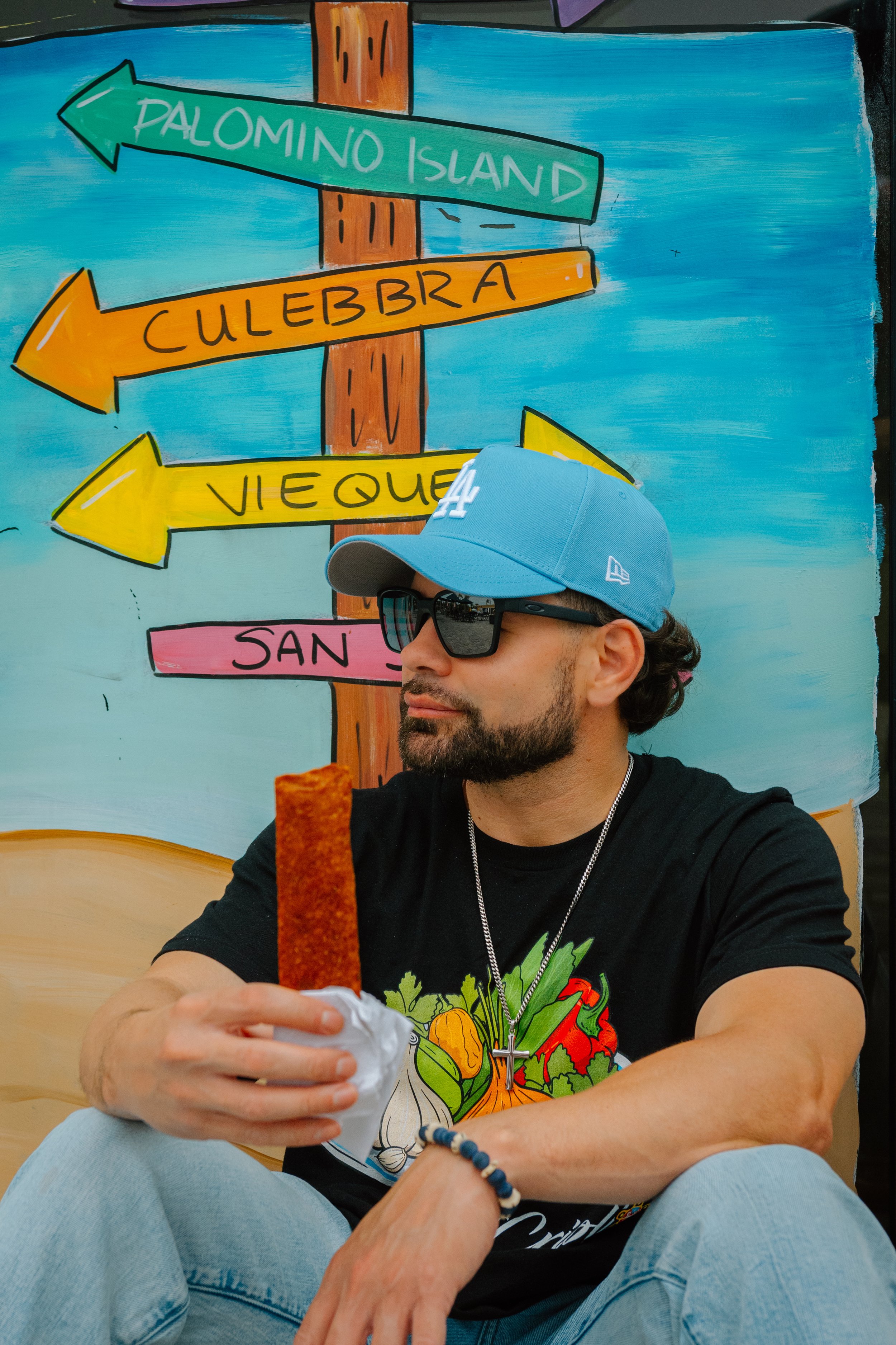 A man wearing sunglasses, a blue cap, and a black T-shirt with a vegetable print, is sitting and holding a churro. Behind him is a colorful hand-painted signpost with arrows pointing to Palomino Island, Culebra, Vieques, and San Juan, on a blue paint