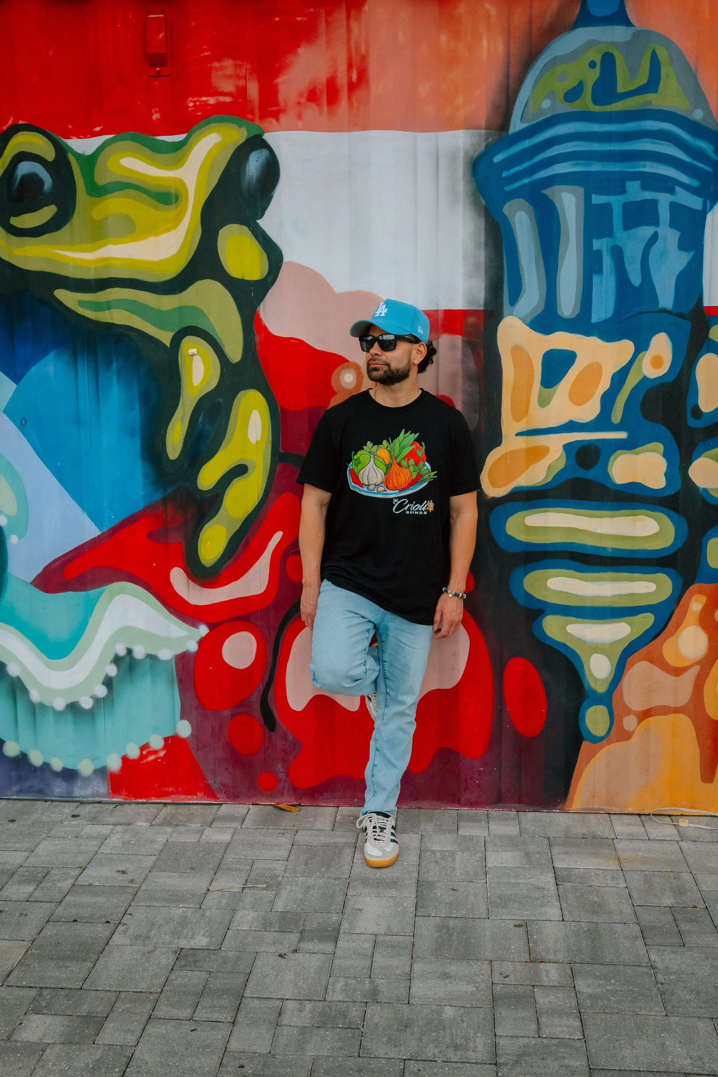 Man standing in front of colorful graffiti wall, wearing black t-shirt with vegetable graphic, light blue jeans, gray sneakers, sunglasses, and a blue Los Angeles Dodgers cap.