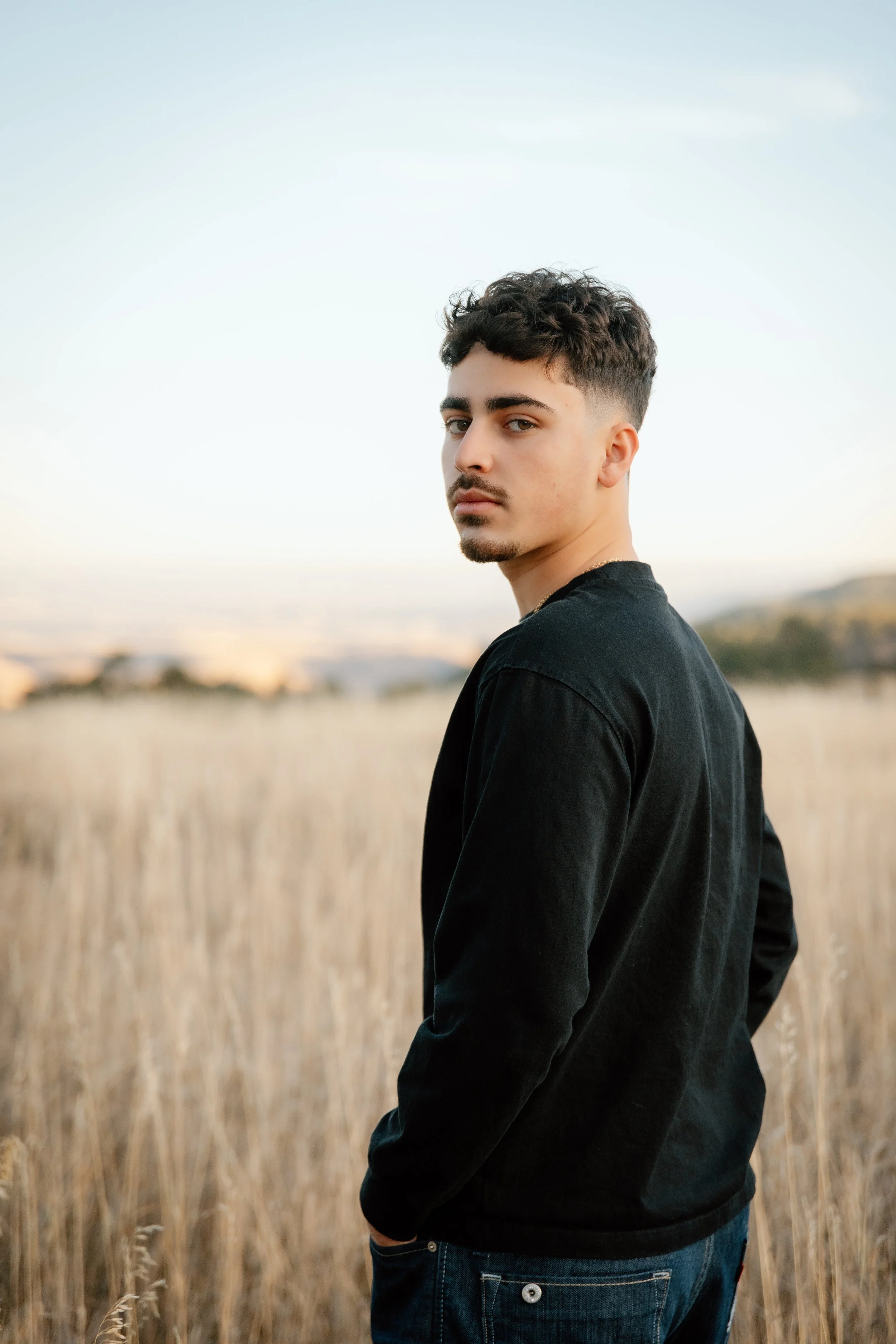 Young man standing in a field of tall dry grass during sunset, looking at the camera with a serious expression, wearing a black long-sleeve shirt and jeans.