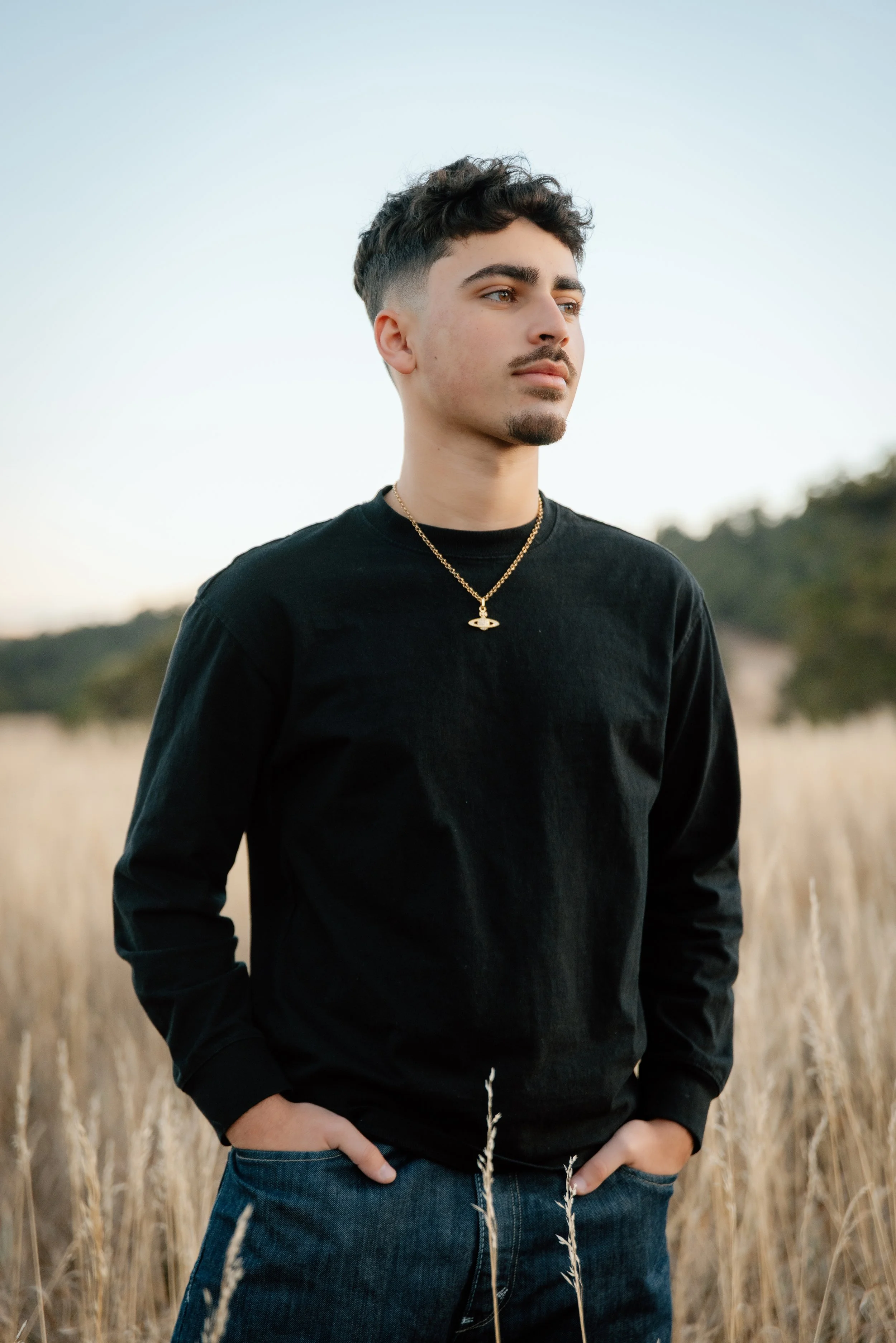 Young man standing in a field of tall, dry grass during sunset, wearing a black long-sleeved shirt and a gold necklace.