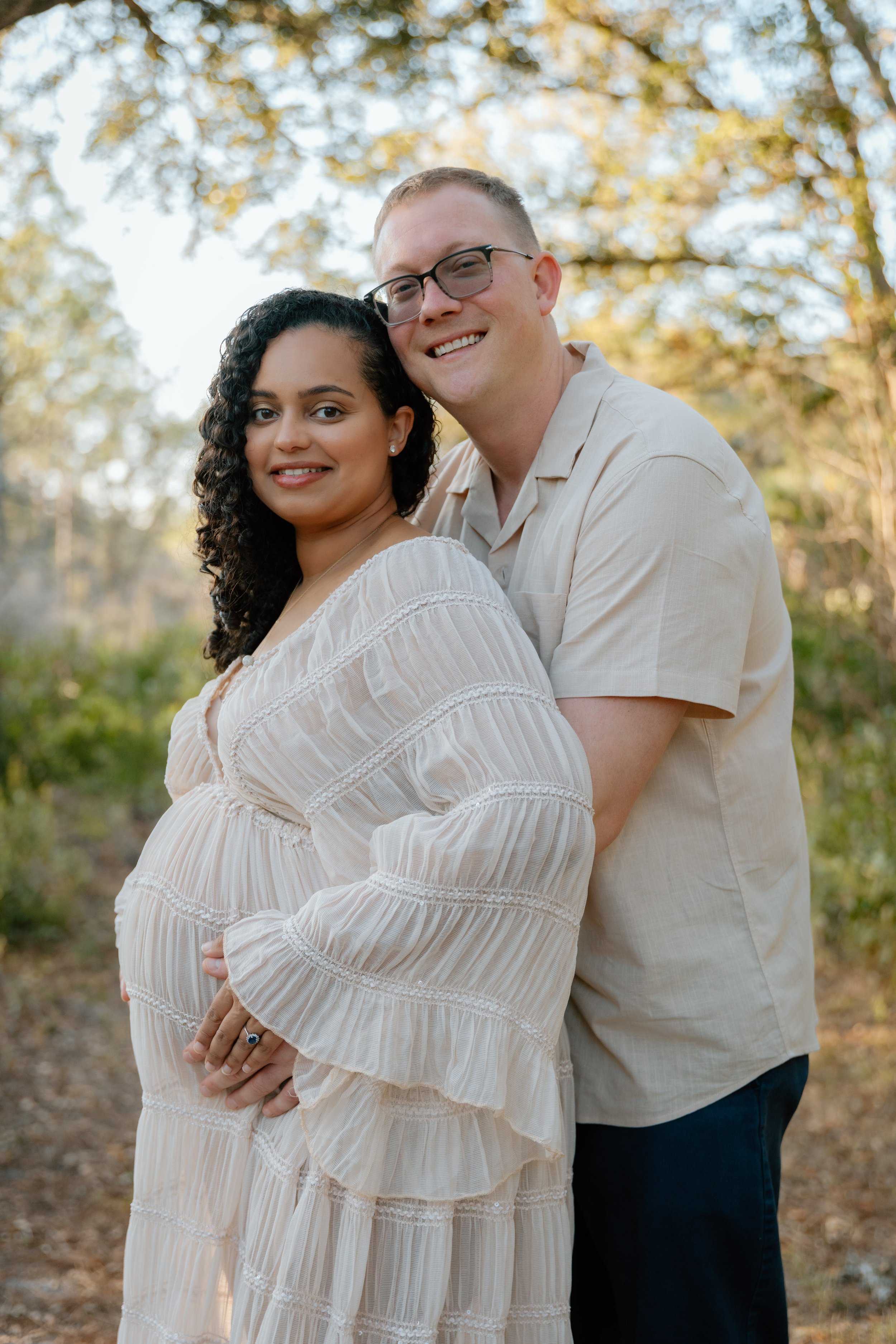 A pregnant woman with curly dark hair wearing a cream-colored dress, standing outdoors, and a man with short light hair and glasses, wearing a beige shirt with rolled-up sleeves, standing behind her and embracing her, in a natural setting with trees 