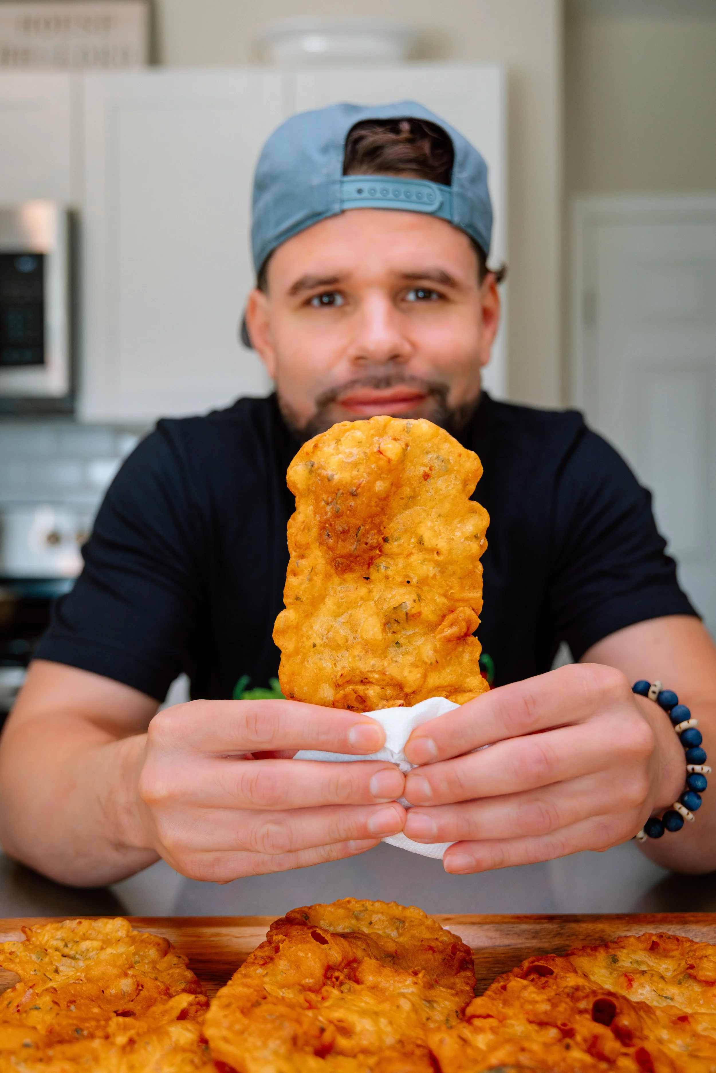 A man wearing a backward baseball cap and a black t-shirt is holding a large, crispy fried food item, possibly a fish fillet or chicken tender, wrapped in a paper towel. There are more fried food items on the wooden surface in front of him.