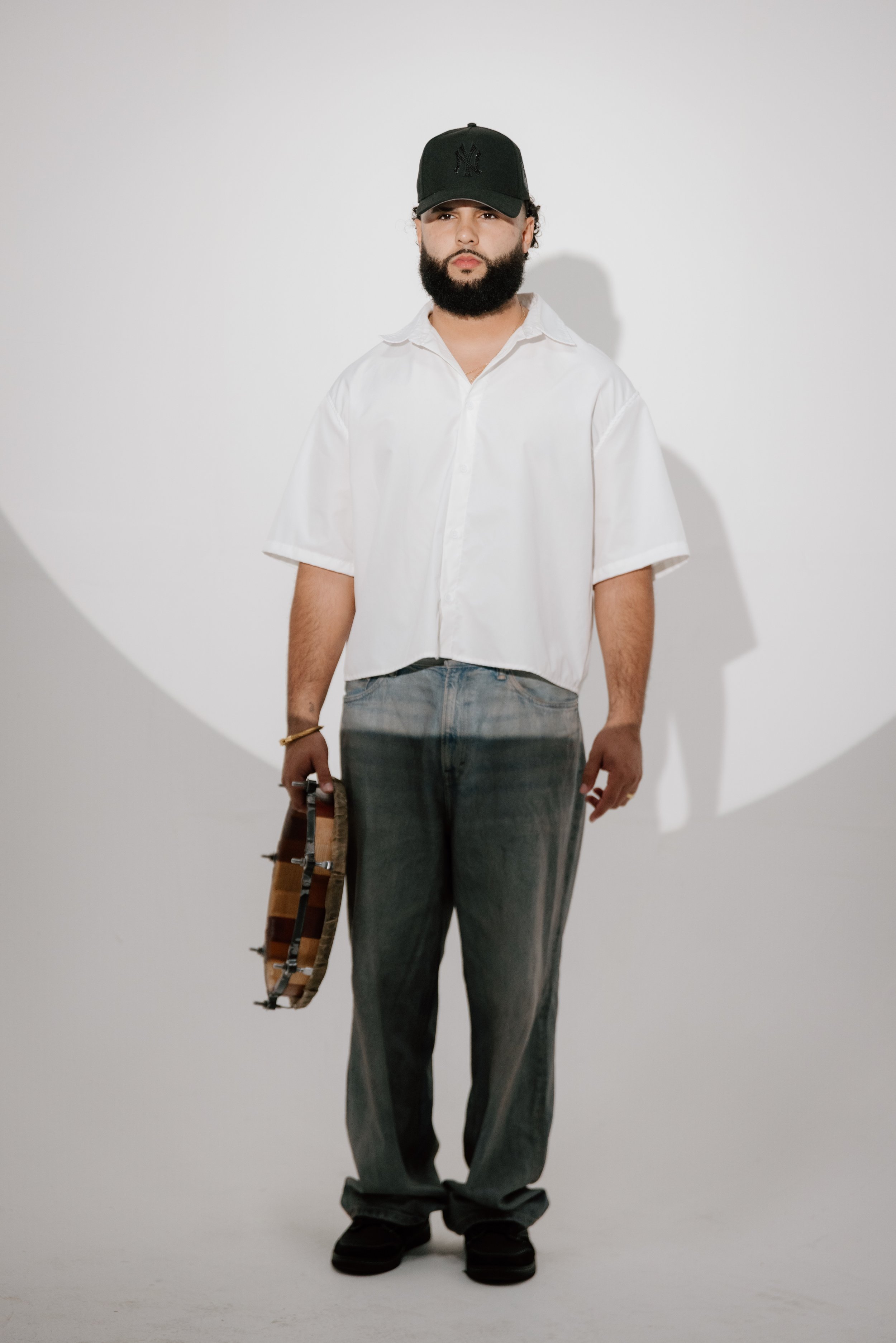 A man with a beard and wearing a black baseball cap, a loose white short-sleeve shirt, baggy jeans, and black shoes, standing against a white background, holding a tambourine in his right hand.