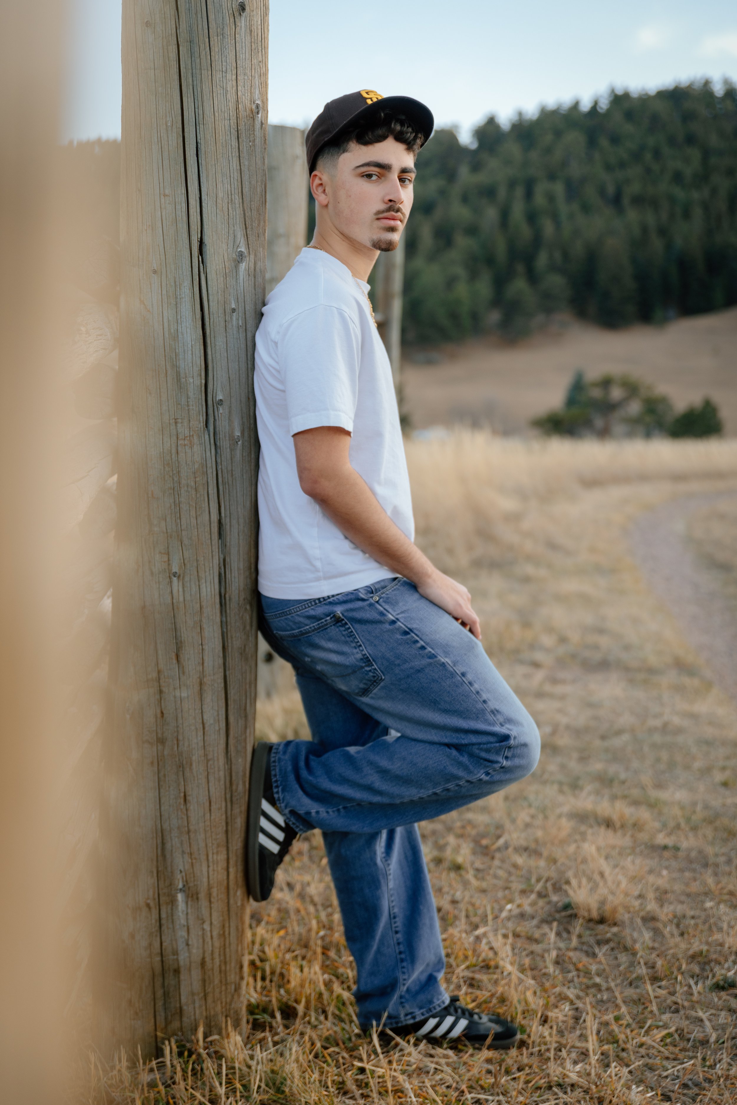 A young man with dark curly hair, wearing a black baseball cap, a white T-shirt, blue jeans, and black sneakers with white stripes, is leaning against a wooden pole outdoors in a rural area with fields and trees in the background.