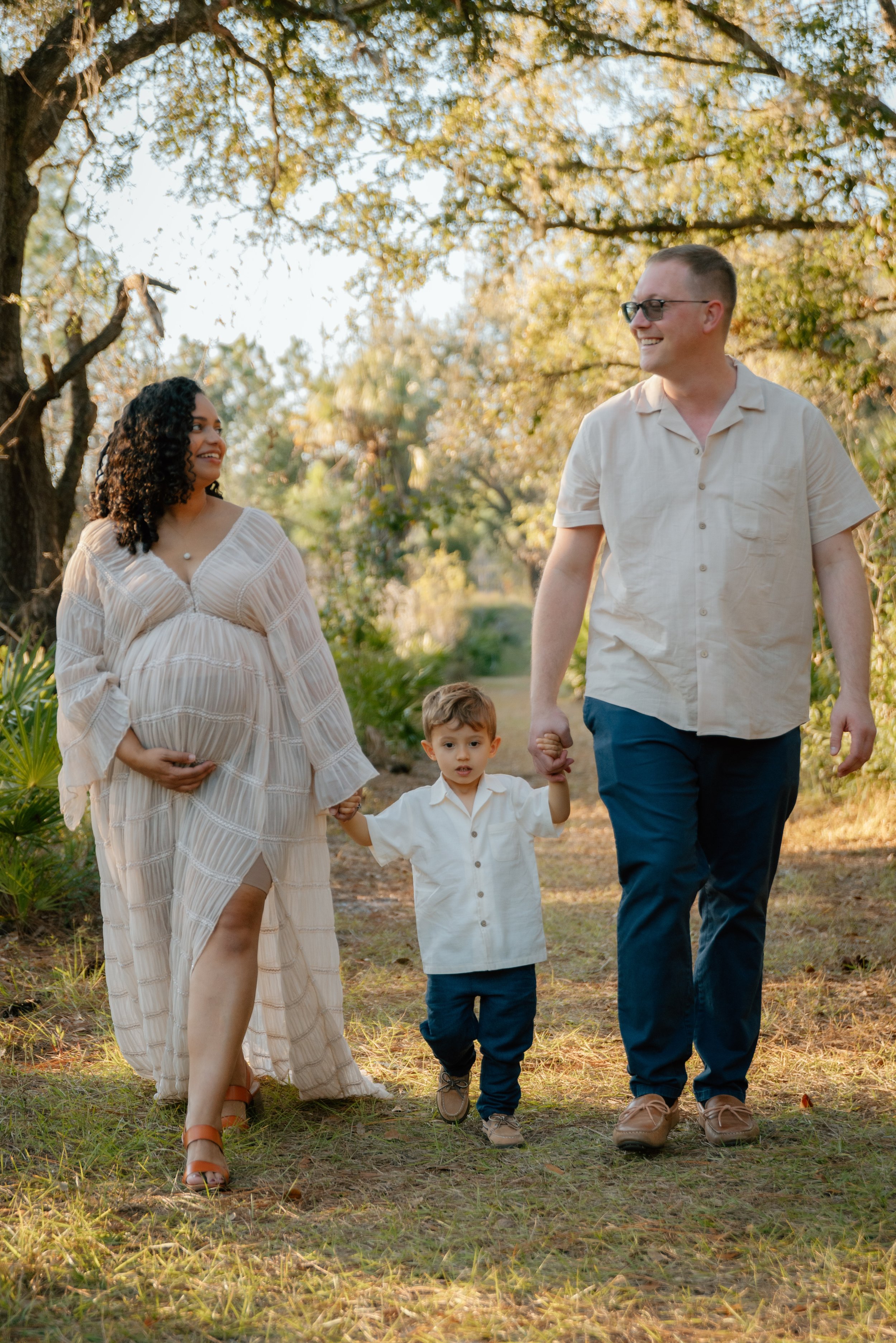 A family of four walking hand in hand outdoors on a sunny day, with trees and greenery in the background. The pregnant woman wears a light-colored dress, the man wears a beige shirt and dark pants, the young boy wears a white shirt and dark pants, an