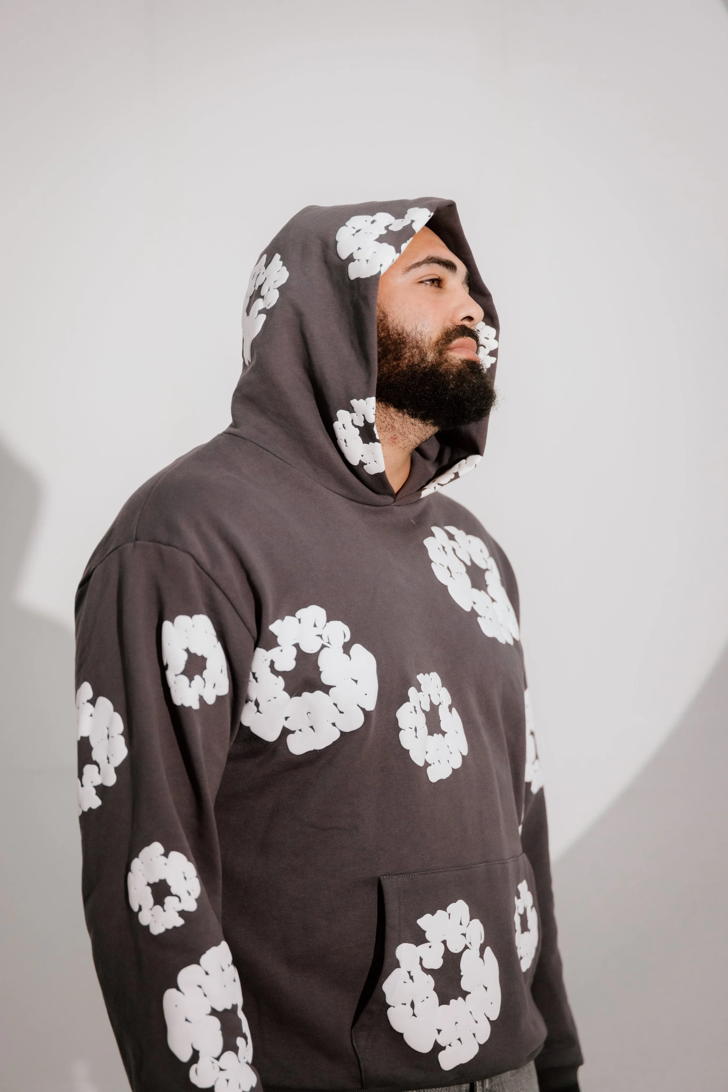 A man with a beard wearing a gray hoodie with white floral patterns, standing against a plain white background.