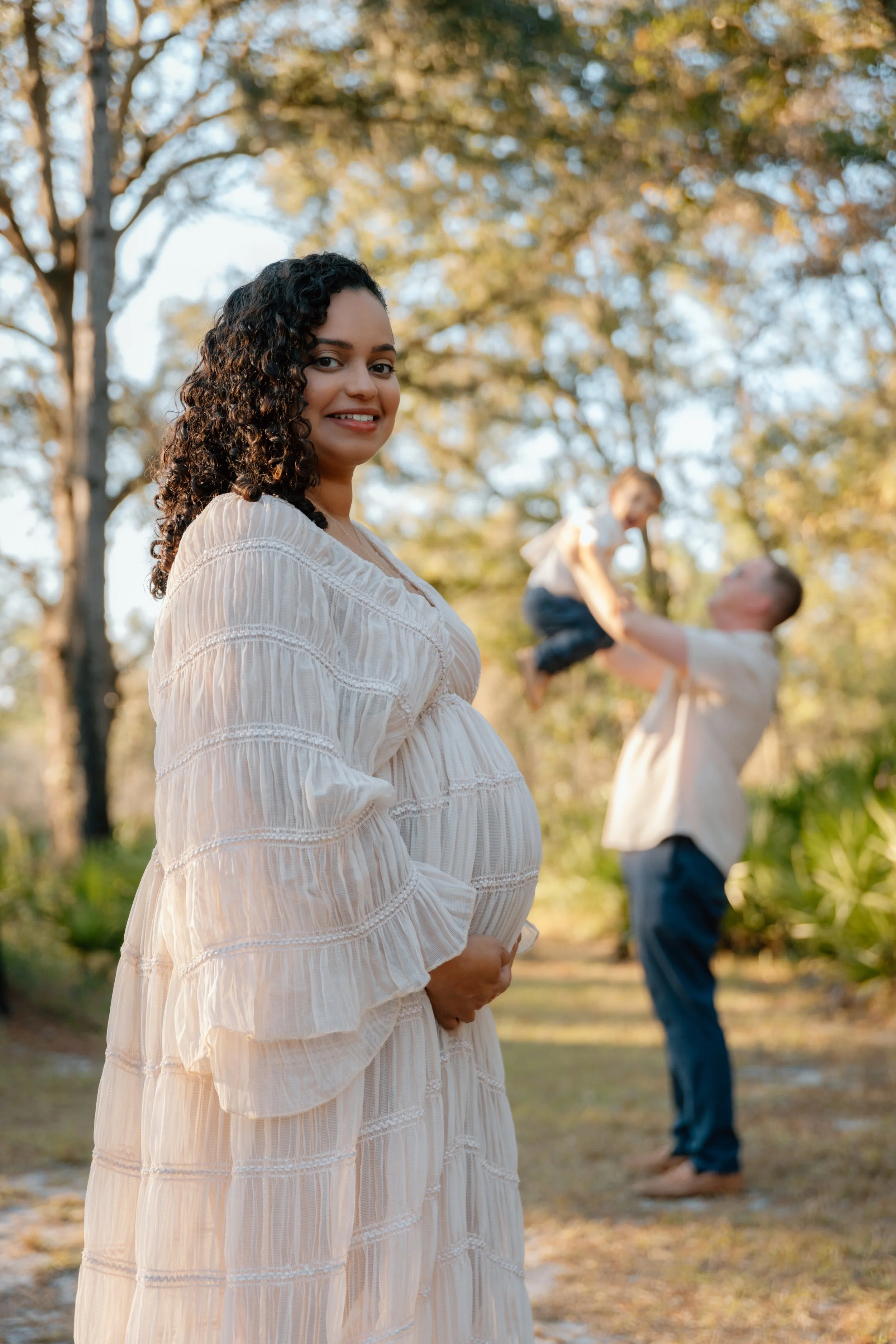 A pregnant woman with curly dark hair smiling outdoors in a white dress, with a man lifting a young girl in the background. The scene is set in a sunny, wooded area.
