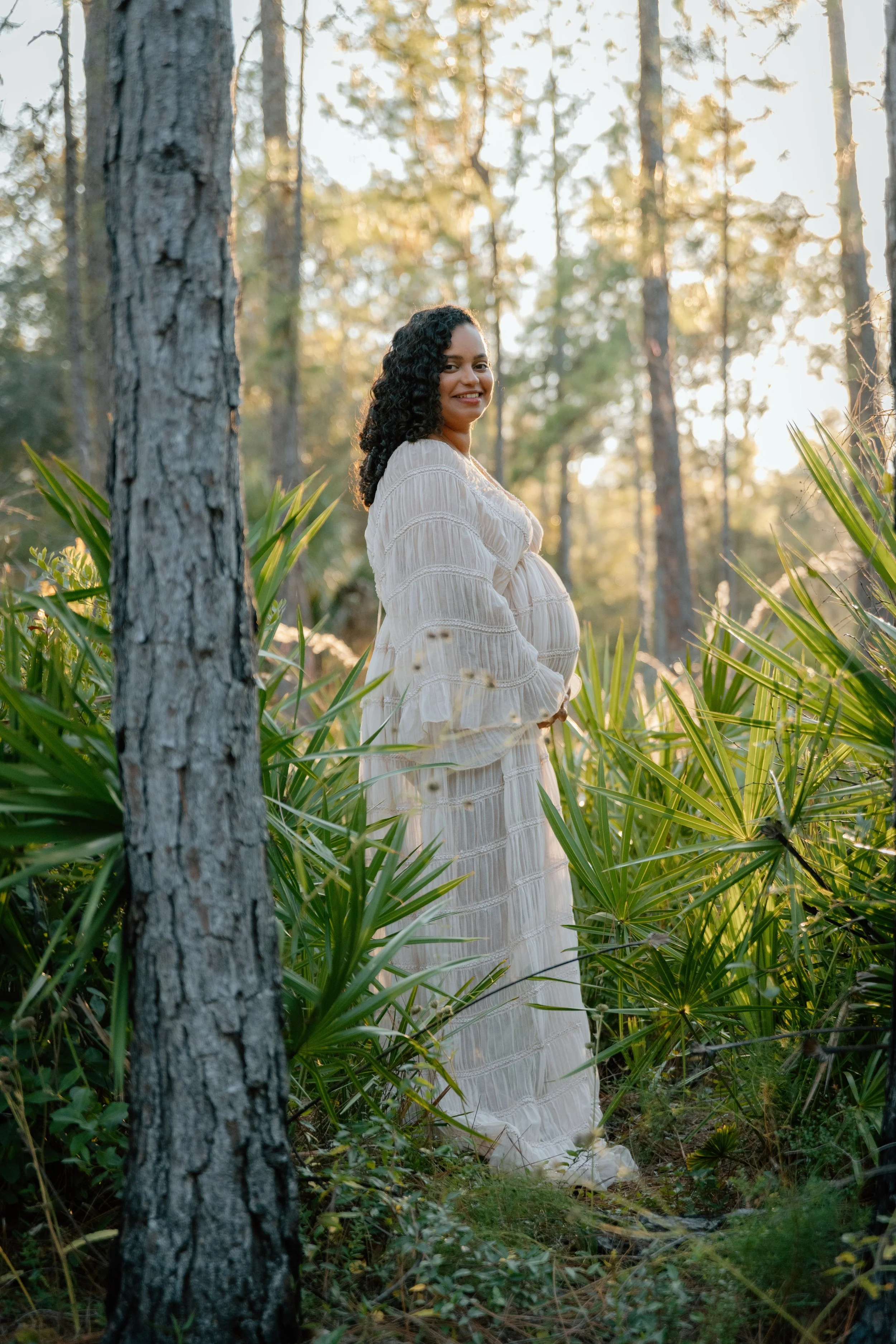 A pregnant woman in a long white dress standing in a sunlit forest with green plants and tall trees, smiling and looking at the camera.