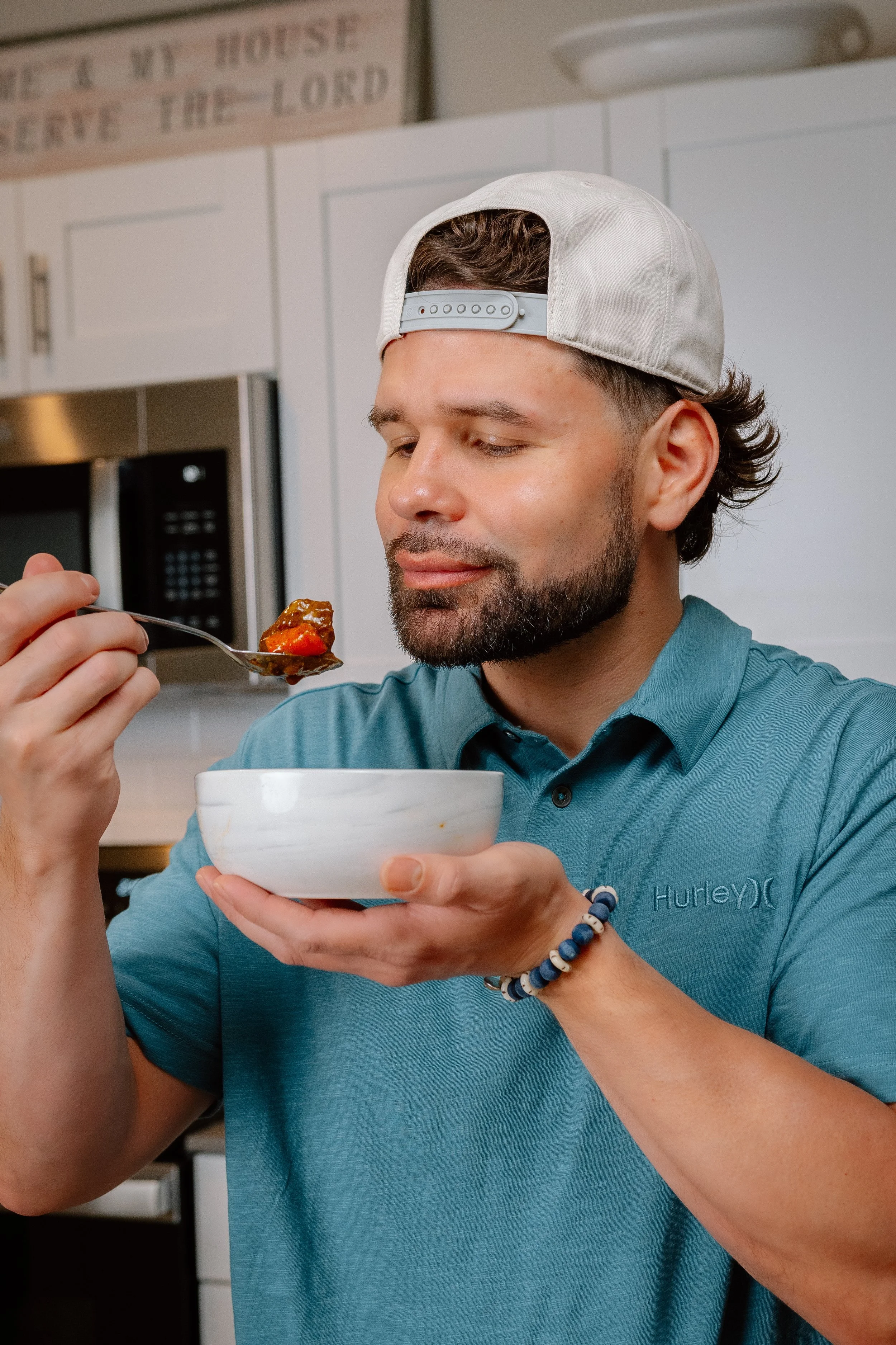 A man with a beard and wearing a blue shirt and white cap eating from a white bowl with a spoon in a kitchen.