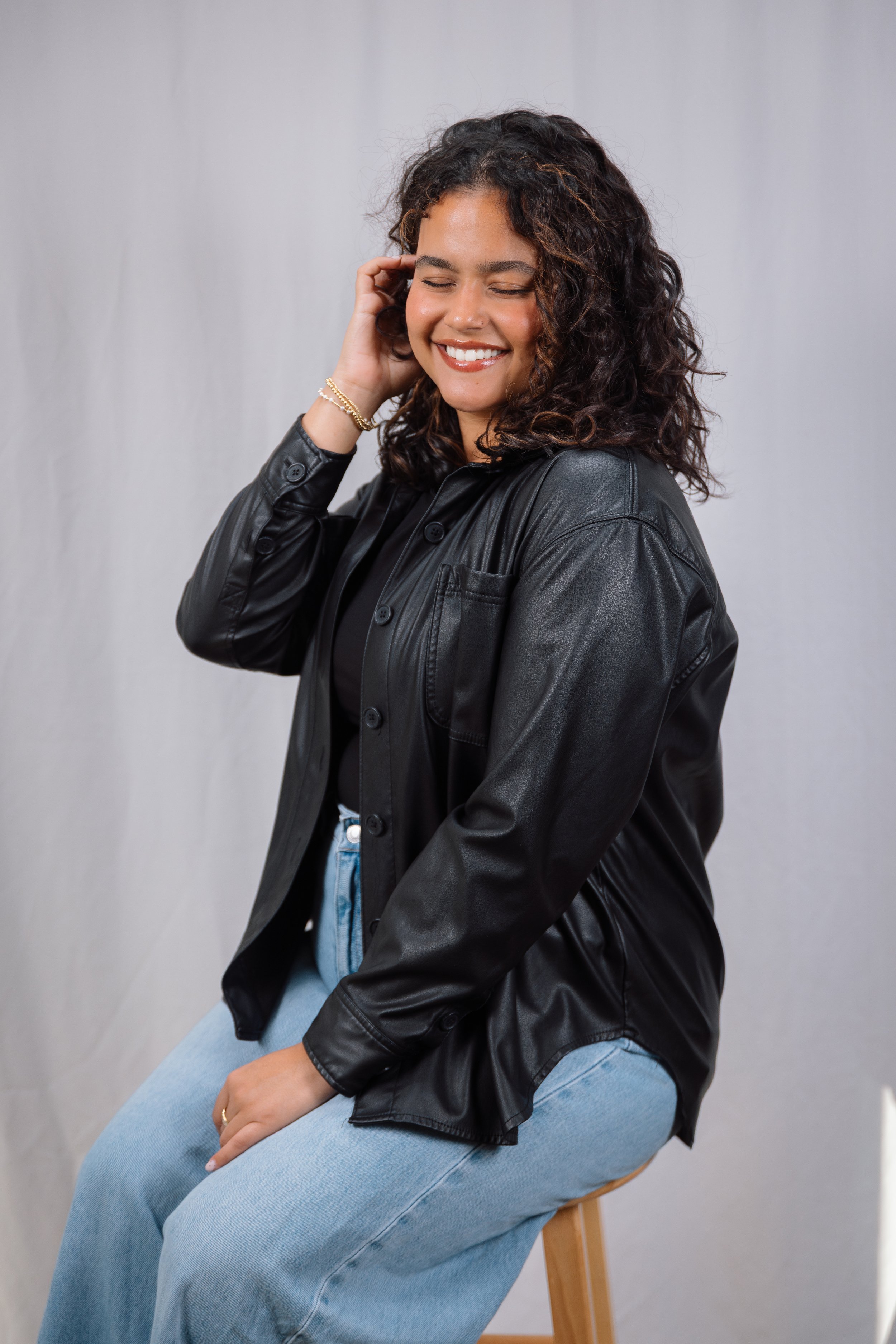 A woman with curly hair smiling, sitting on a wooden stool, wearing a black leather jacket and light blue jeans.