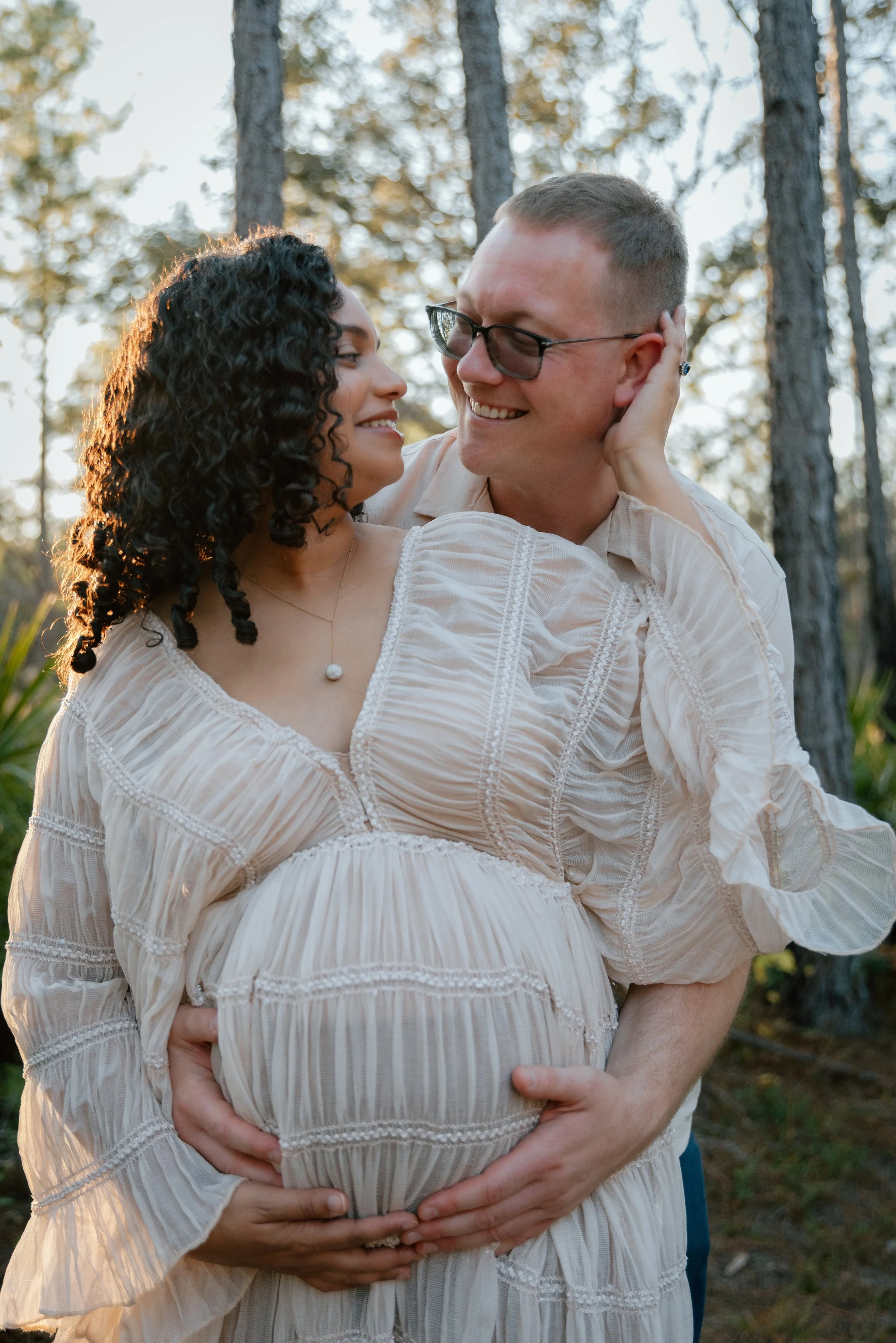 A pregnant woman and a man smiling and embracing in a forest during sunset, with the woman touching her belly and the man holding her midsection.