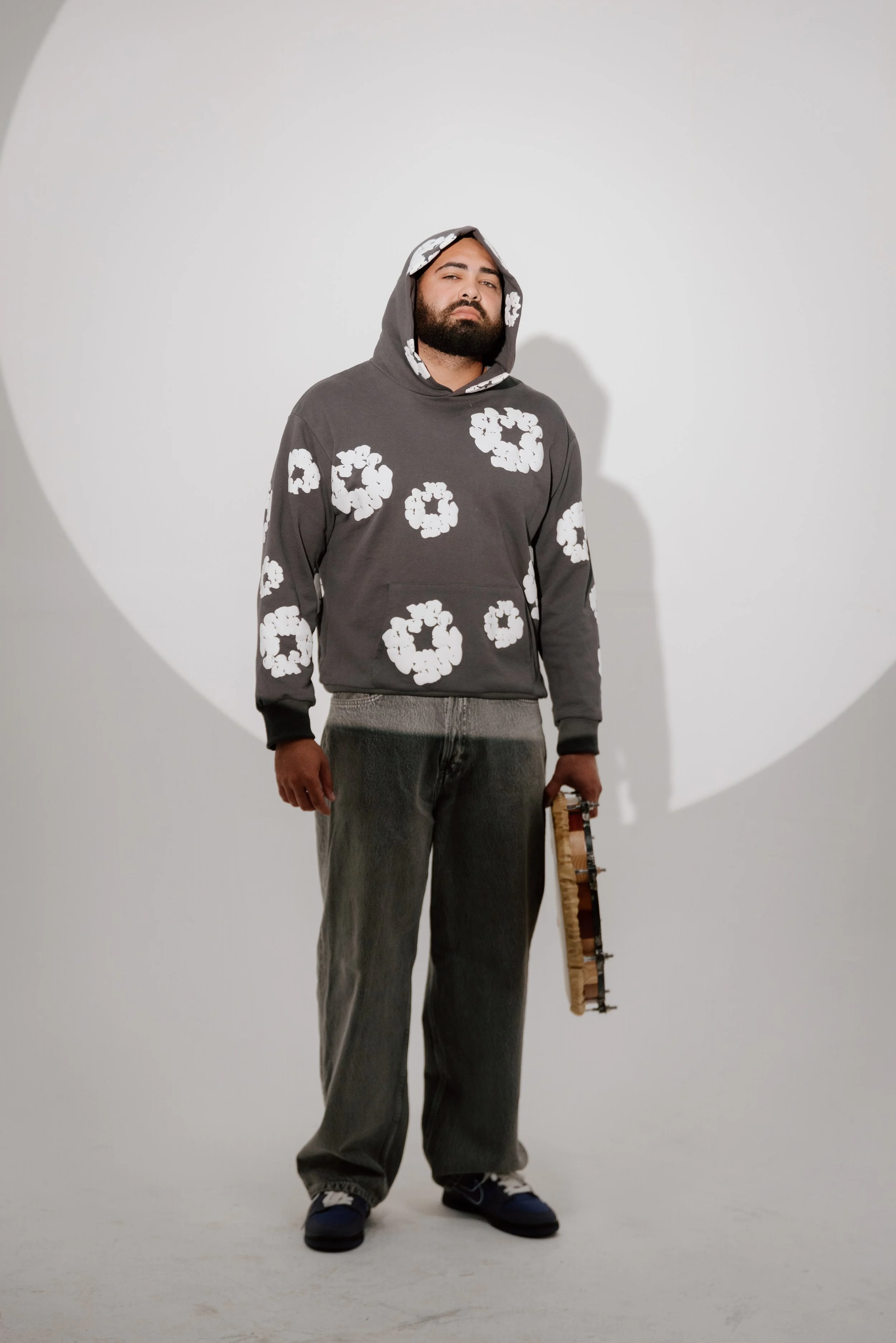 A man standing in front of a plain white background, wearing a dark hoodie with white flower patterns, gray jeans, and shoes, holding a small object in his right hand.
