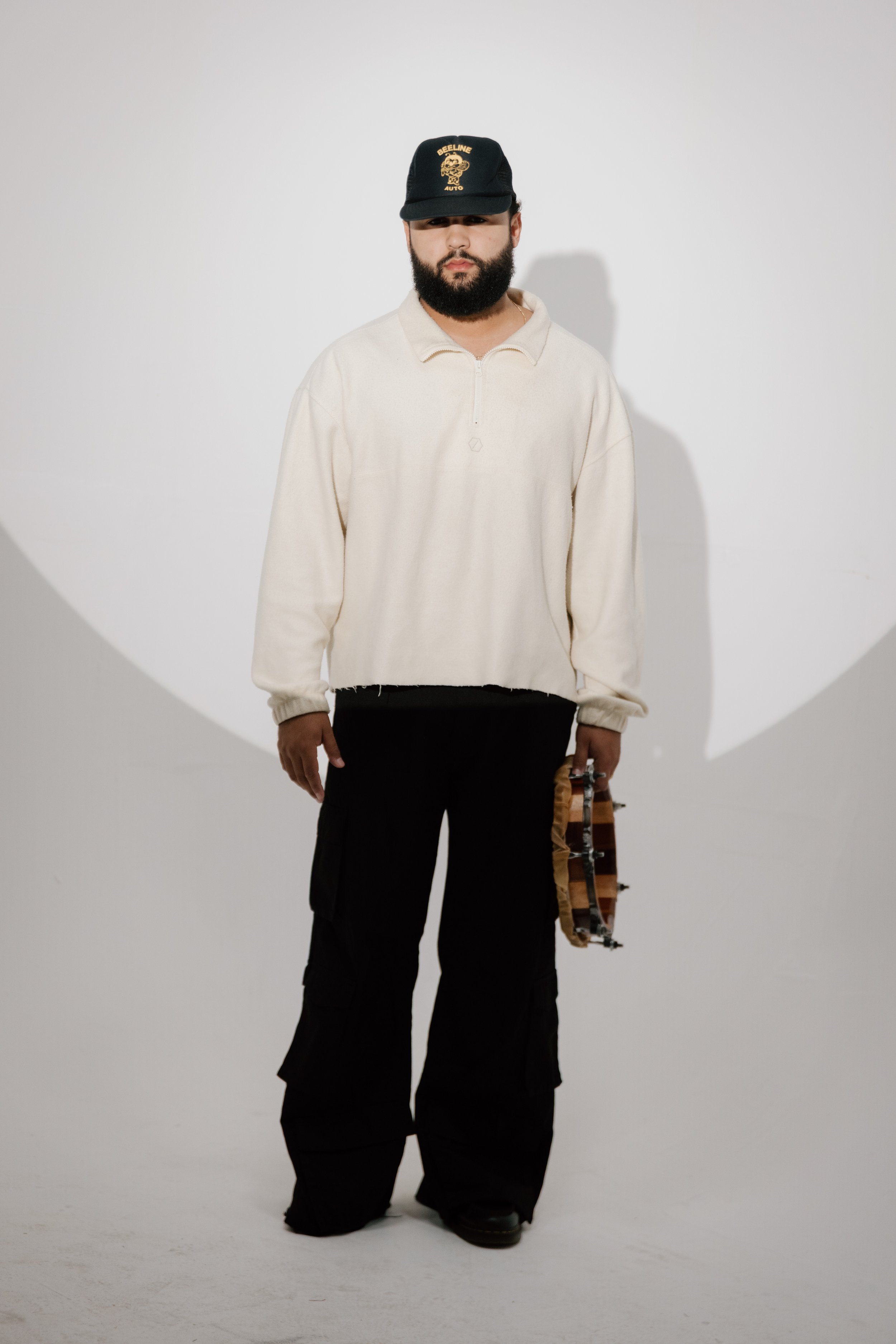 A man with a beard wearing a black cap, a light-colored oversized pullover, black cargo pants, and holding a skateboard, standing against a white background with a spotlight shadow.