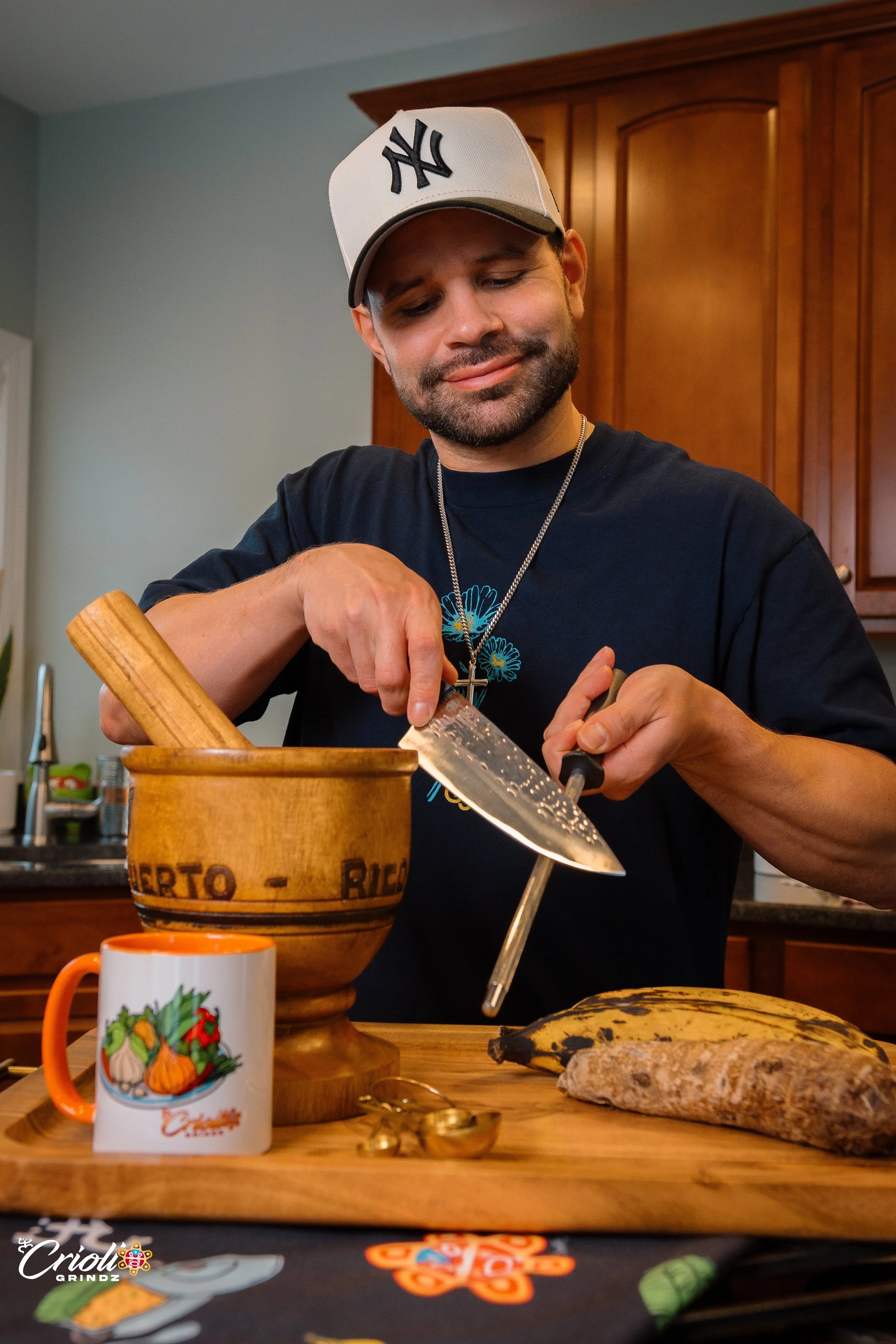 A man wearing a New York Yankees cap and a black t-shirt with blue flower design is chopping food in a kitchen, with a wooden bowl, bananas, a root vegetable, and a Halloween-themed mug on the counter.