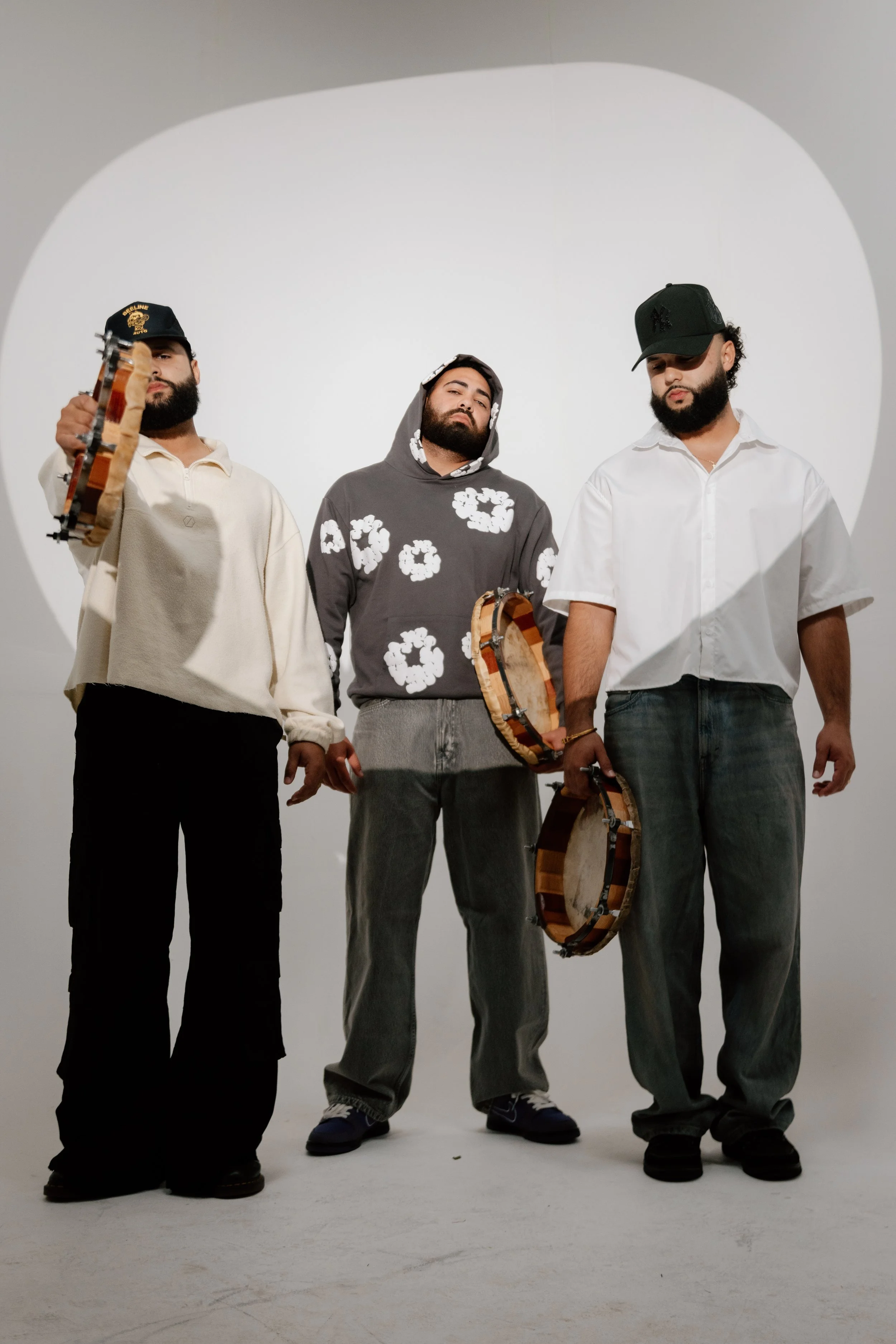 Three men standing together in a photography studio with a white backdrop, holding small drums, and wearing casual streetwear including hats and hoodies.