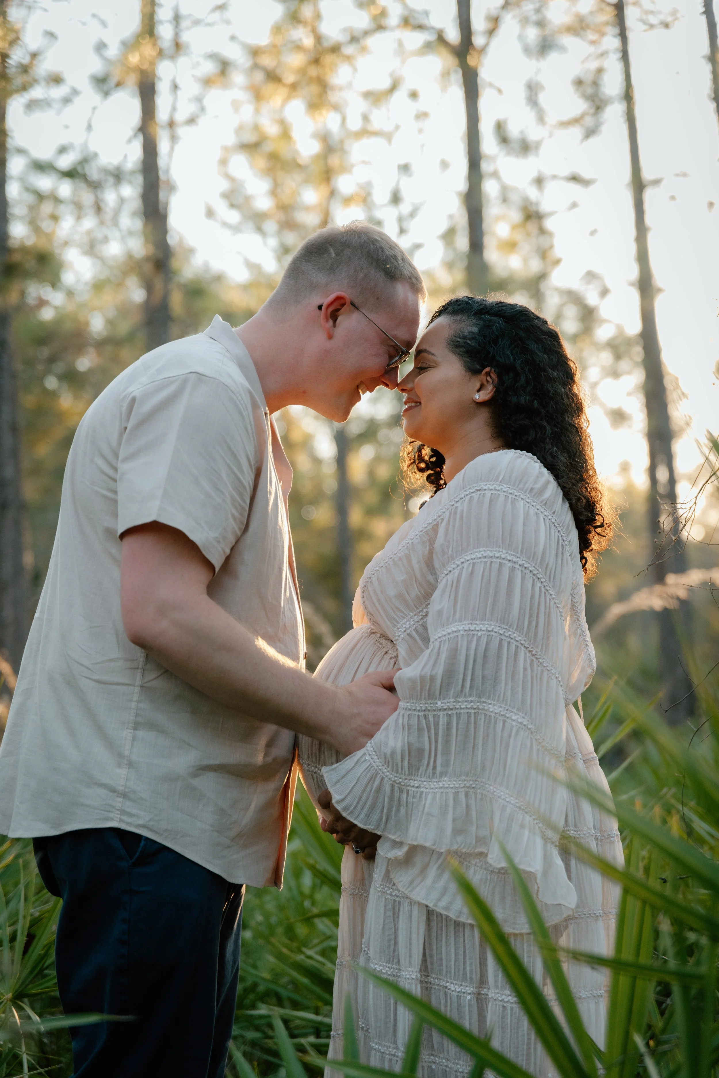 A couple standing close together in a forest at sunset, touching foreheads and holding hands with a pregnant woman wearing a white dress and a man in a beige shirt.