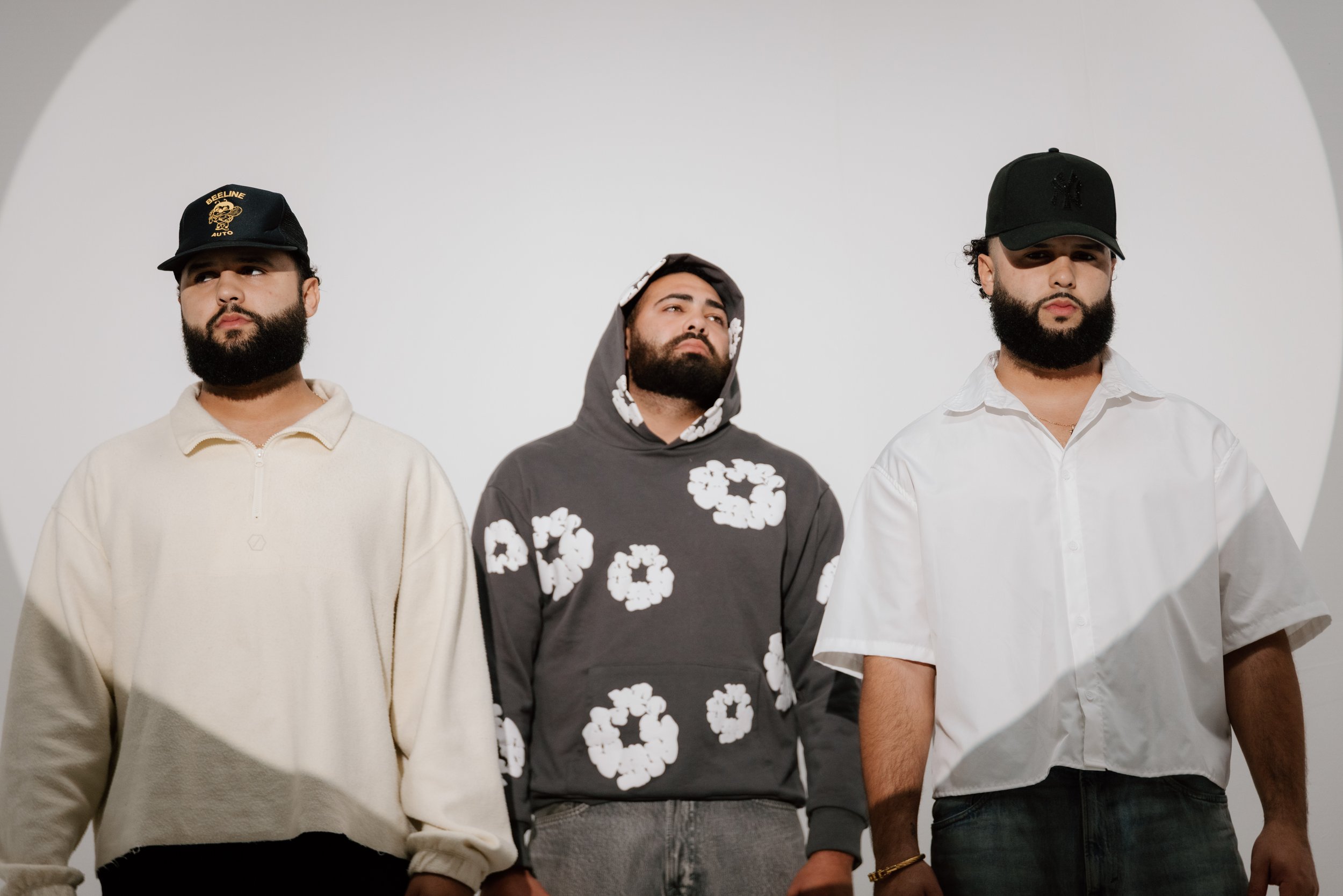 Three young men standing side by side against a plain white background, each wearing casual streetwear and hats.