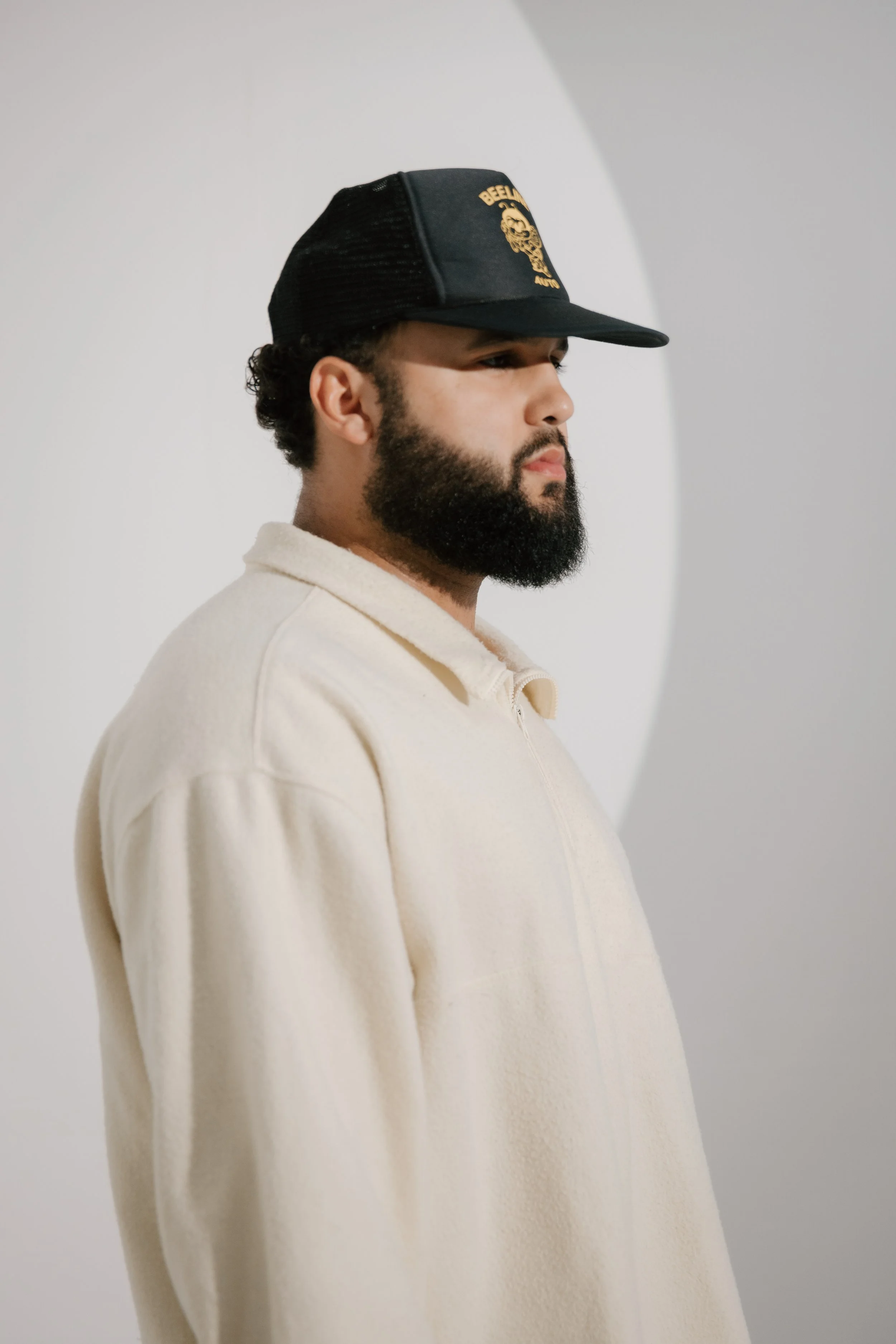 Profile of a man with a beard wearing a beige fleece jacket and a black trucker hat with a gold logo.
