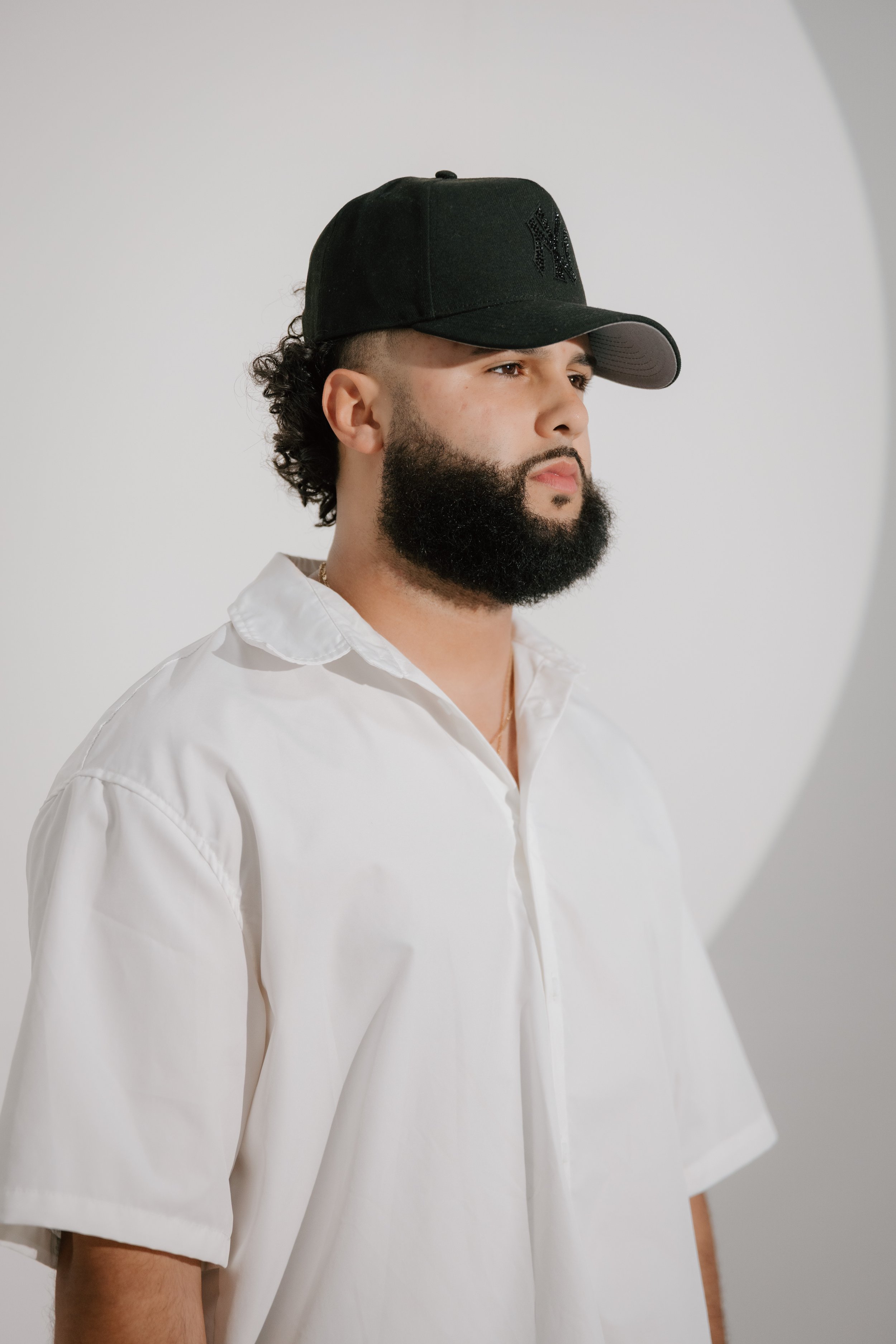 A man with a beard wearing a white shirt and a black baseball cap, standing against a plain background.