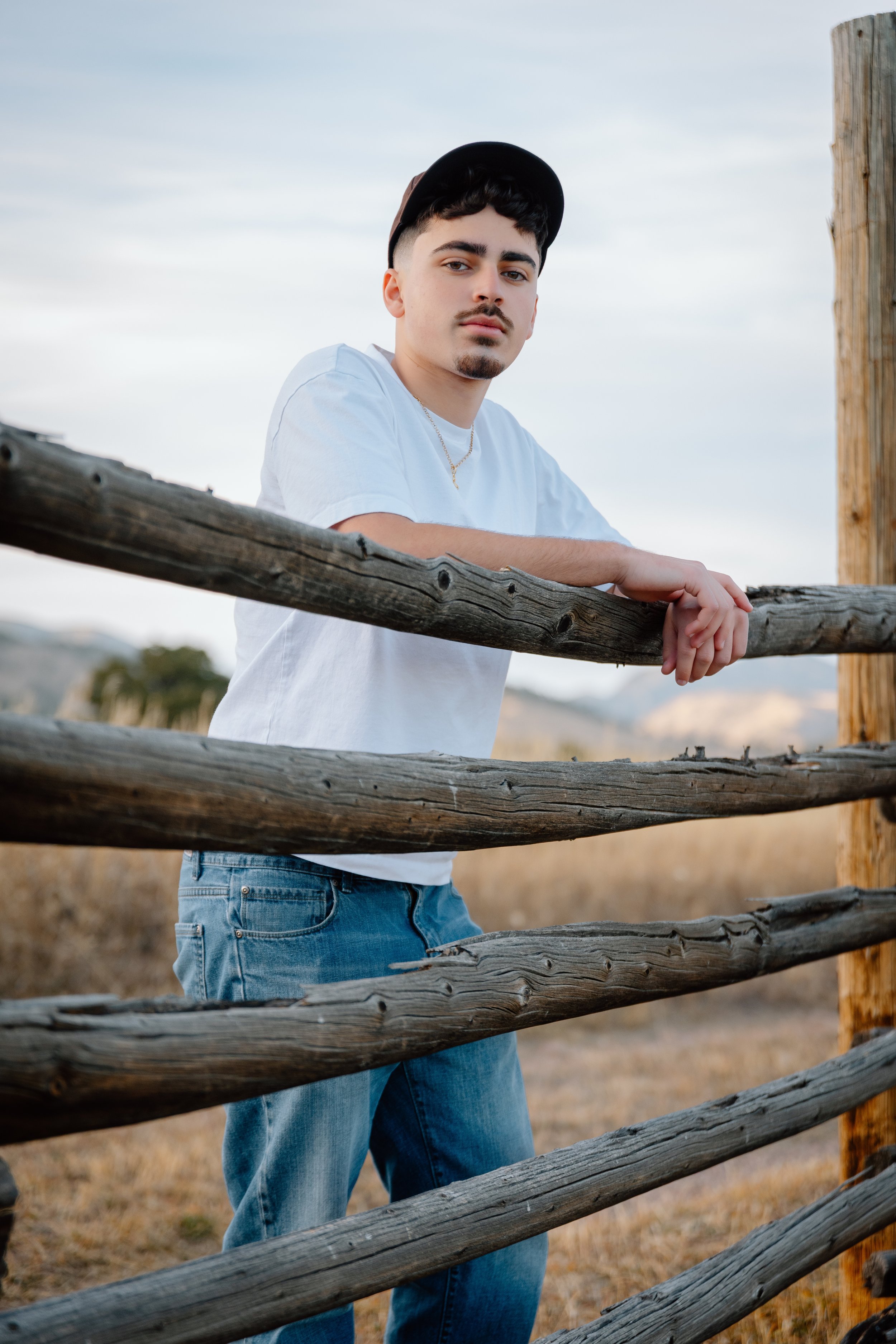Young man leaning on rustic wooden fence outdoors, wearing a white t-shirt, jeans, and a black cap, with a neutral expression, landscape background with hills and clear sky.