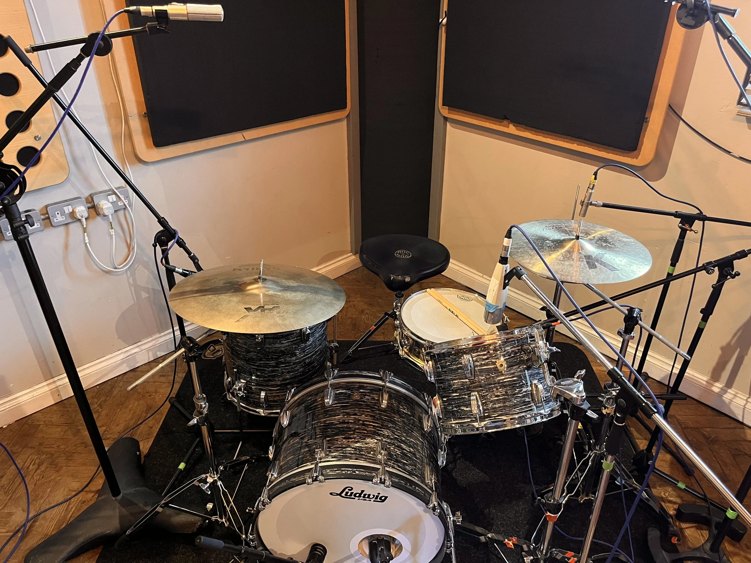 A drum set in a recording studio with microphones positioned around it, surrounded by soundproofing panels on the walls.