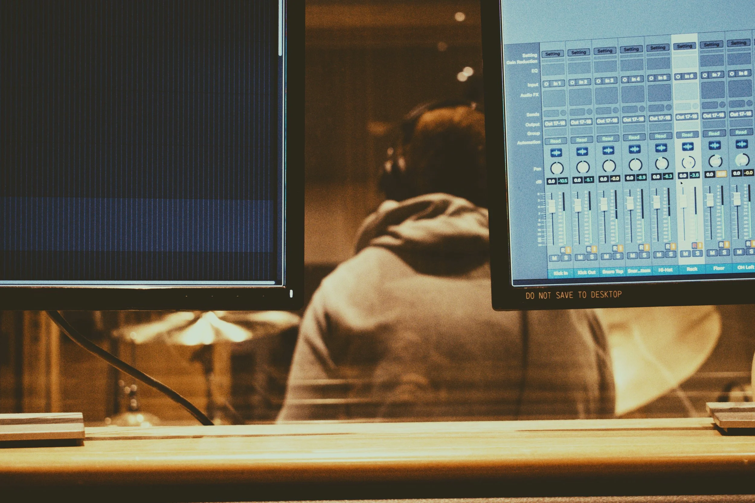 Recording studio with two computer monitors displaying audio editing software, a person wearing headphones working in the background, and a desk surface in the foreground. Recording for Picture Day, a Brighton based indie band.