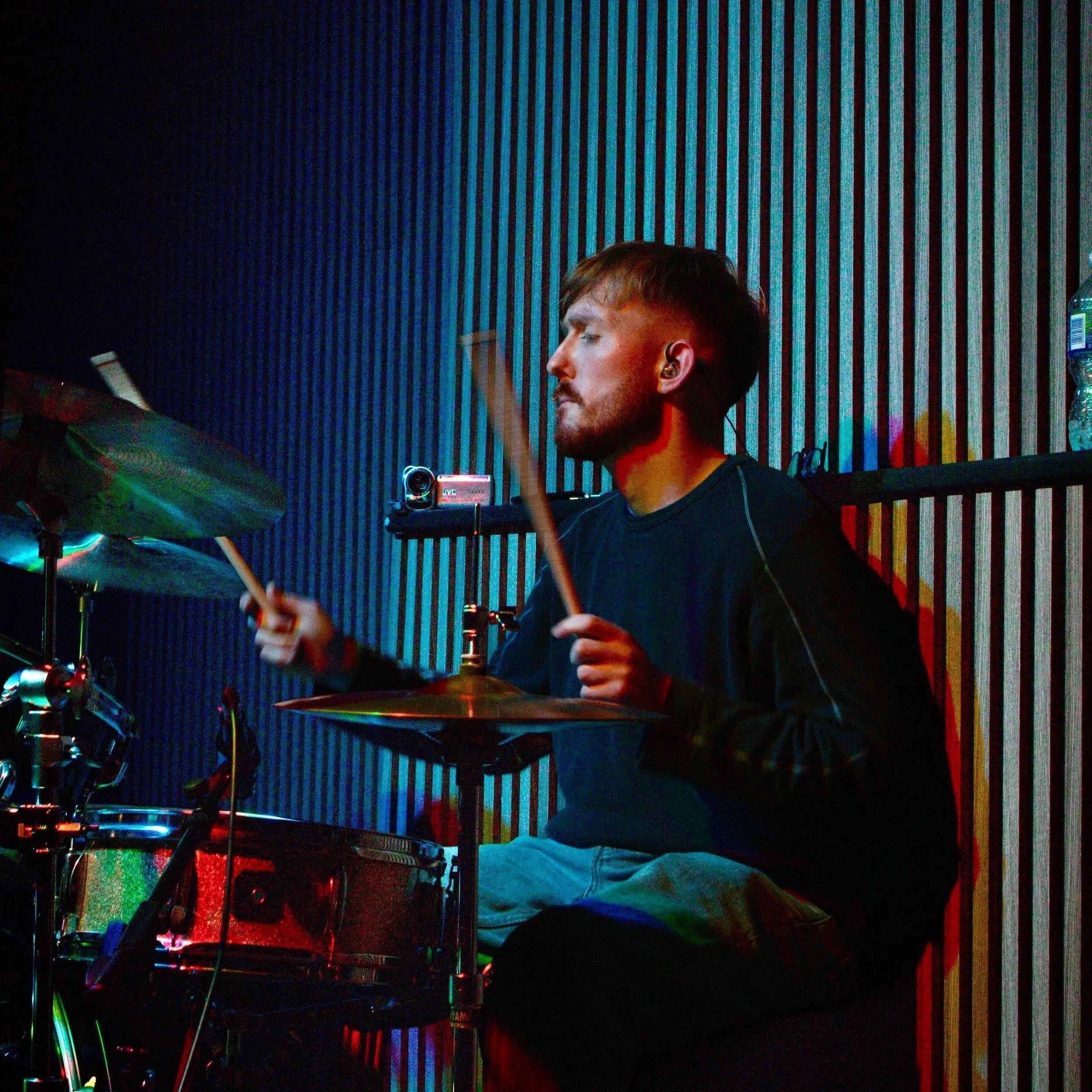 A man with earphones playing drums in a room with colorful lighting and a striped wall.