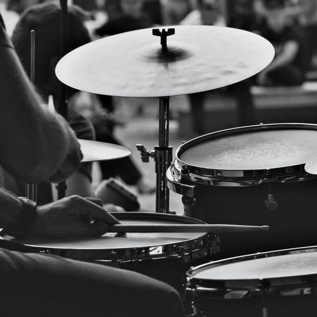 Black and white photo of a drummer playing a drum set with cymbals, in an outdoor setting with blurred people in the background.