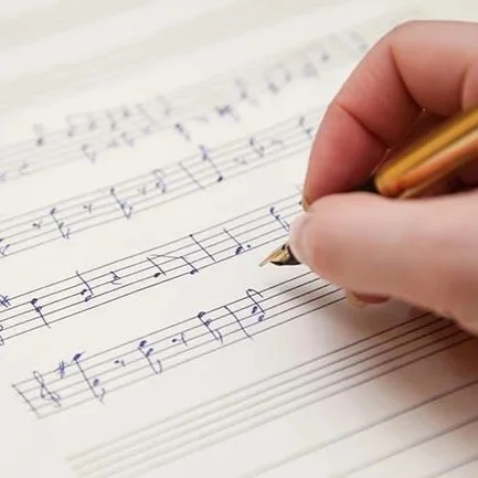 Person writing musical notes on sheet music with a gold pen.