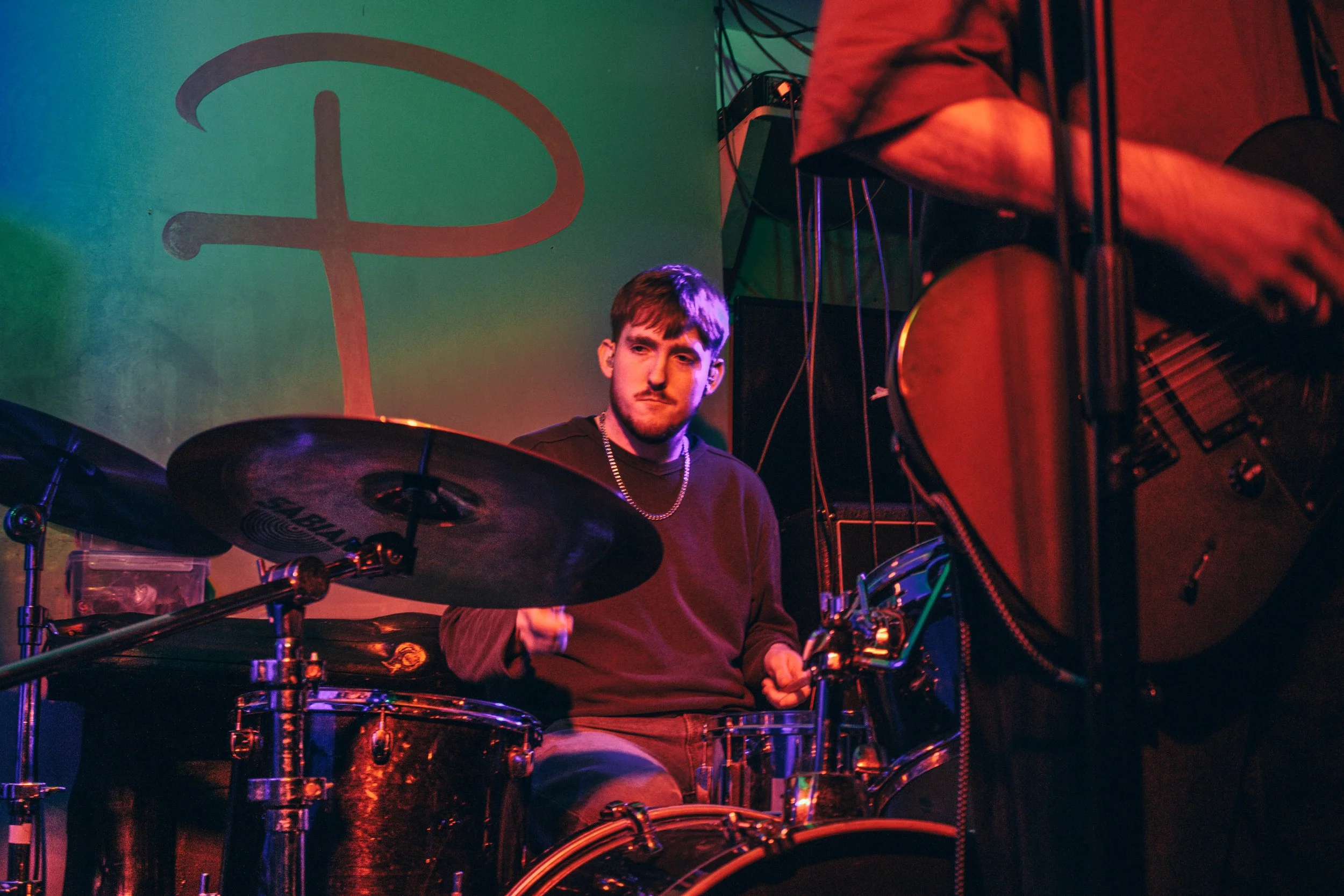 A young man playing drums and another person playing guitar on stage with a colourful background. He is playing with Sleepflower, a Brighton based alternative band. The venue is Pipeline, Brighton, which is a pub that has a music venue above it
